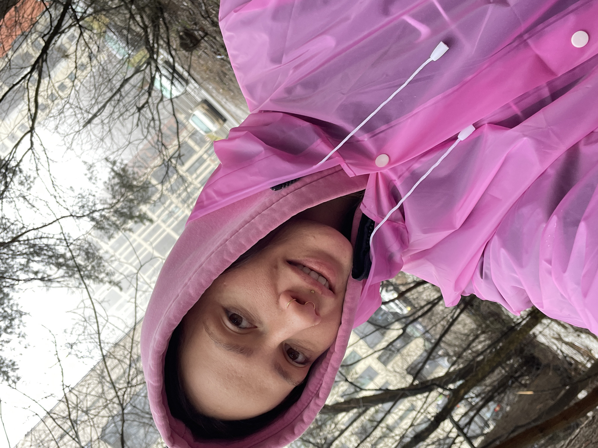 A woman wearing a pink raincoat with a hood poses for a selfie-style photograph. The background consists of bare tree branches and a tall building, suggesting an urban environment. The photograph is taken at an angle, as the buildings appear to be in the sky. The woman appears to be fair-skinned, with a nose ring and a slight smile. The image provides no indication it was taken in Izhevsk, Russia.