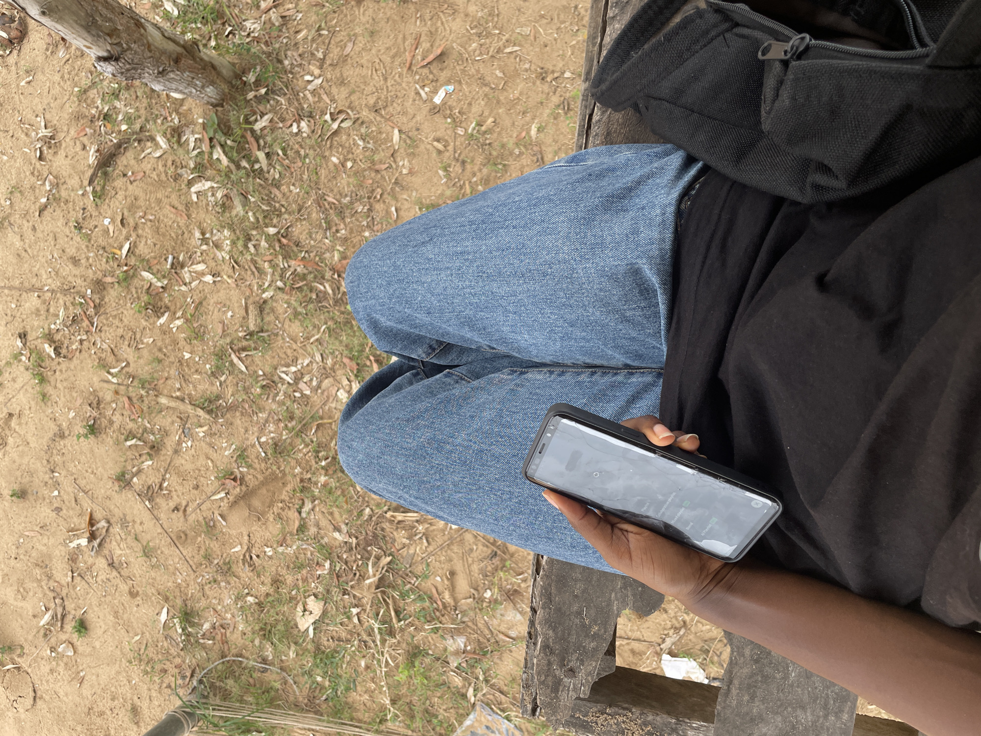 A first-person perspective shot shows a person sitting on a wooden bench outdoors. The person is wearing blue jeans and a black t-shirt. They are holding a dark-colored smartphone in their left hand, which displays a list of music playlists. The ground is covered with dry leaves, sand, and sparse green grass. A tree trunk is visible on the right side of the frame. The scene appears to be a casual outdoor setting, possibly a park or a quiet area. The provided crops offer closer views of the ground texture, the smartphone's screen, and the person's clothing and hand. There is no specific information or visual cues in the image to identify the location as Iyafin, Nigeria.