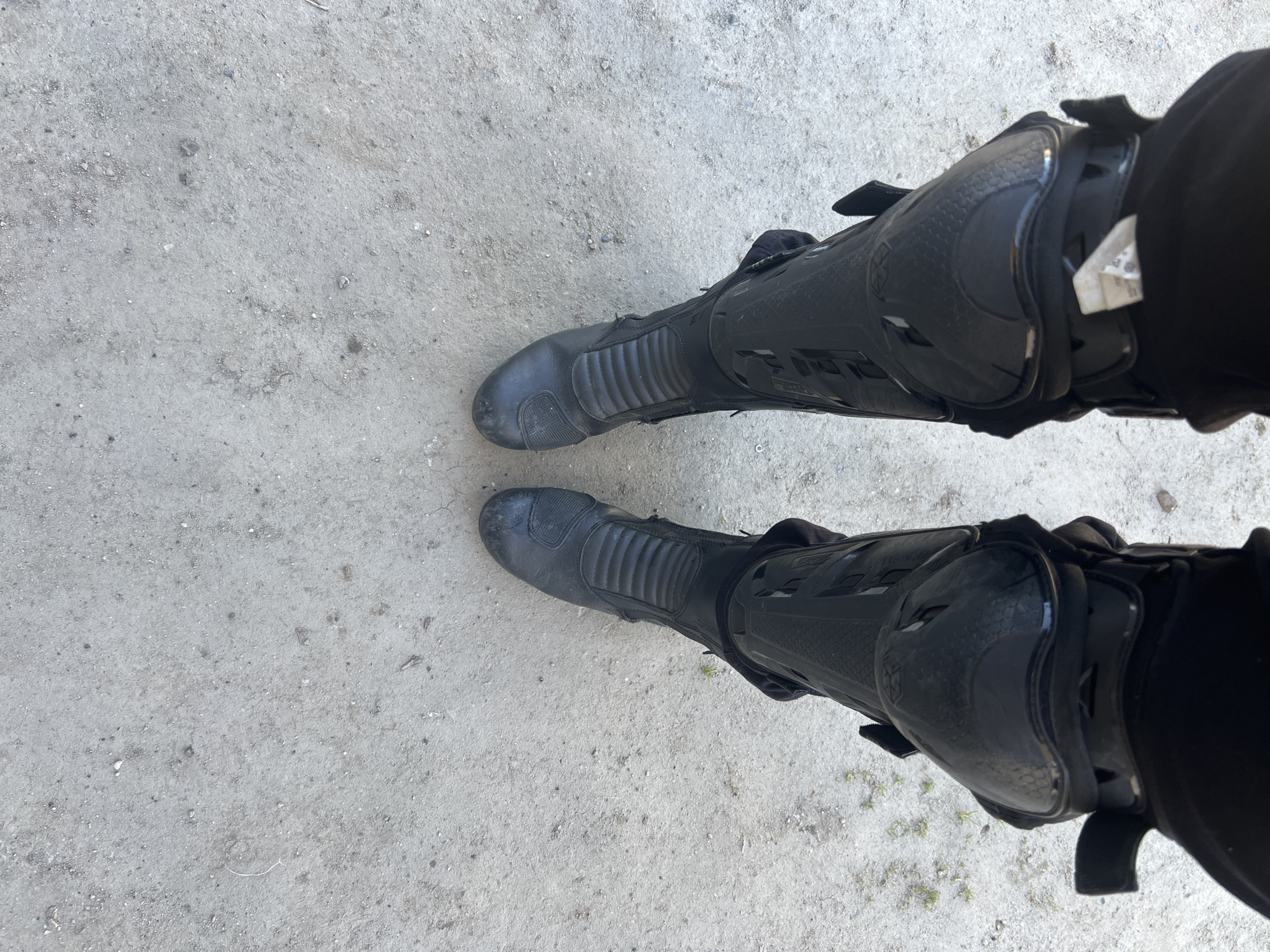 The image shows a first-person perspective of two legs wearing black motorcycle boots standing on a concrete surface. The boots appear to be slightly worn and dusty. The concrete surface is light grey with small pebbles and imperfections visible. The location is not identifiable as Cherliany, Ukraine from the image alone.