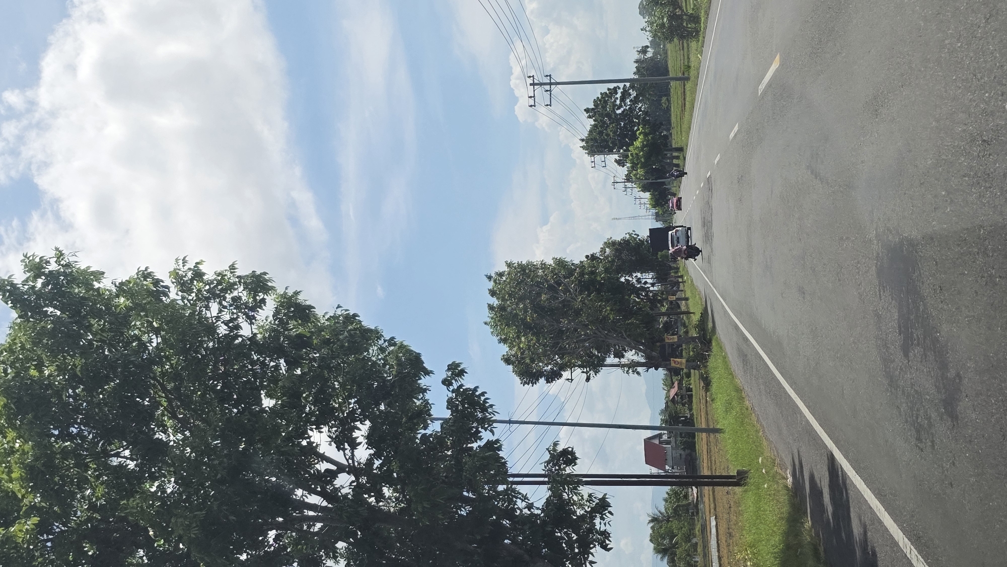 The image depicts a road scene in Kampungjumpa, Indonesia, showcasing a two-lane asphalt road with a white line separating the lanes. Trees line the left side of the road. Power lines stretch across the sky. A vehicle and a motorcycle are visible on the road. The sky is blue with scattered clouds. Grass and a house are visible on the roadside.