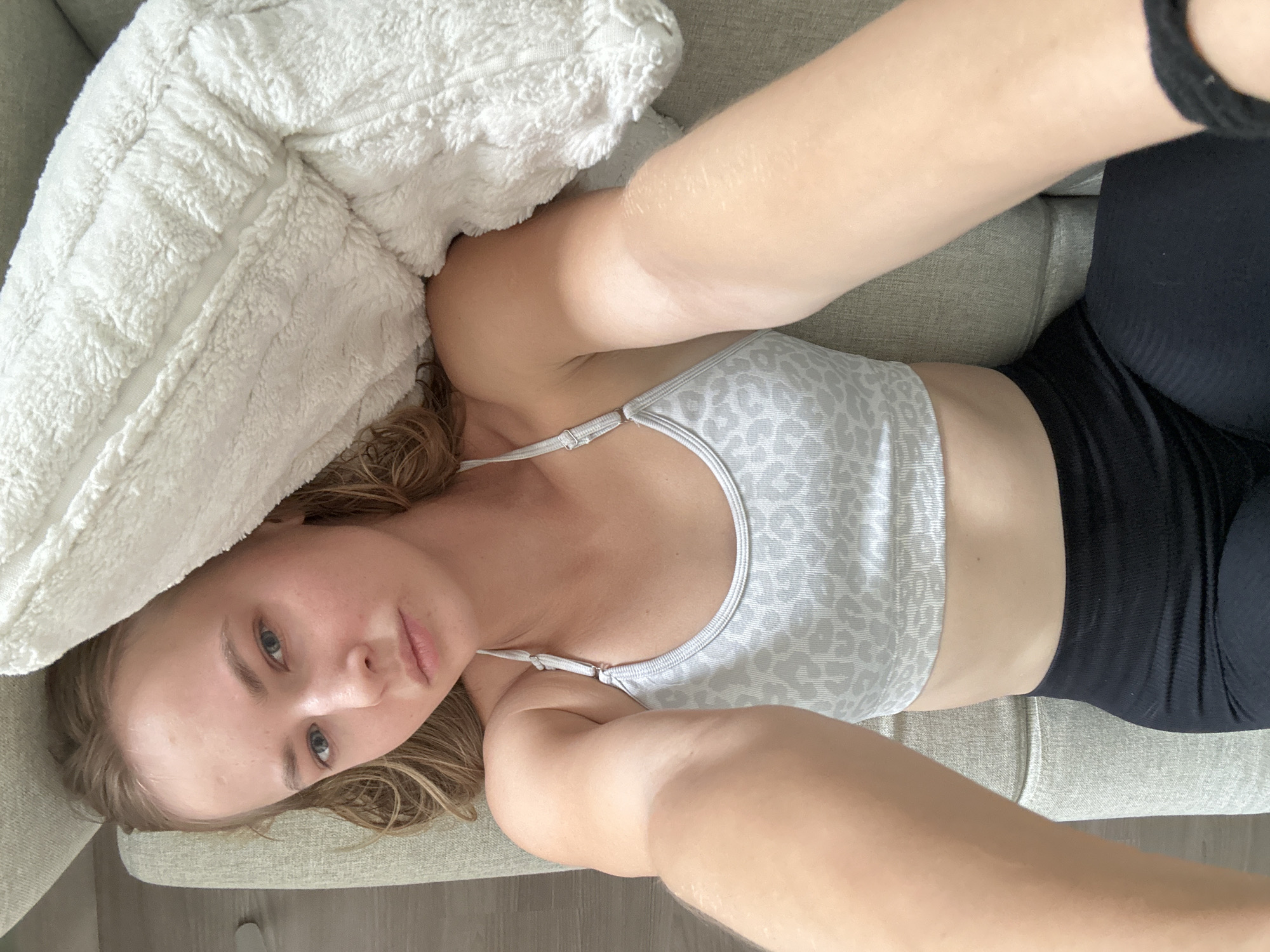 A young woman is lying on a sofa, her head resting on a fluffy white pillow. She is wearing a grey leopard-print sports bra and black leggings. Her eyes are open and she is looking at the camera, with a neutral expression. The room appears to be a living room, with a sofa and a wooden floor visible. The lighting suggests it is daytime. The image is a selfie, taken from a high angle.