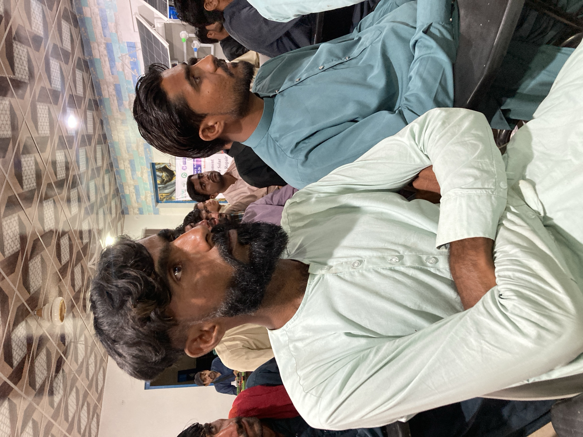 The image shows a group of men sitting indoors, likely in a meeting or presentation. The men are dressed in traditional South Asian clothing, specifically shalwar kameez. The man closest to the camera has a dark beard and is wearing a light green shalwar kameez. He is sitting with his arms crossed. Several other men are visible in the background, wearing different colors of shalwar kameez. The background suggests an office or community center. There's a patterned tile floor and a sign on the wall, which could provide context about the location or purpose of the gathering. The location in the city is specified as Unknown, Unknown.