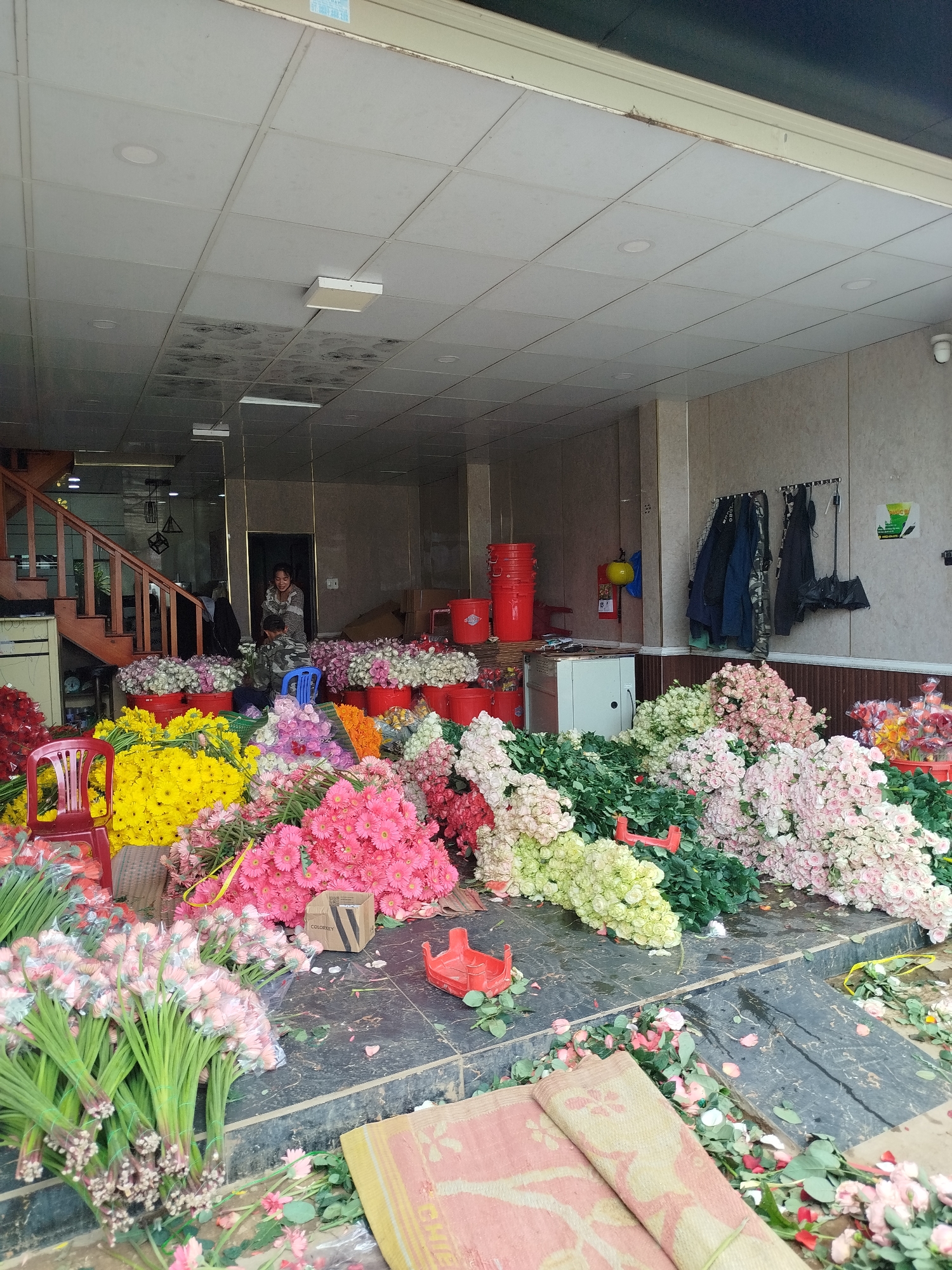 The scene depicts the interior of what appears to be a flower shop located in Cam Ly, Đà Lạt, Vietnam. The shop is overflowing with flowers of various colors including red, yellow, pink, and white. Some flowers are in buckets, while others are scattered on the floor. Two people, a woman and a child, are visible in the background standing near a doorway and next to a set of stairs. In the background, red buckets are stacked near a white refrigerator and a yellow fire extinguisher. Clothes and an umbrella are hanging on the wall to the right. Two rolled-up mats are in the foreground. The floor is littered with flower petals and stems. The overall impression is of a bustling flower business with a wide assortment of flowers.