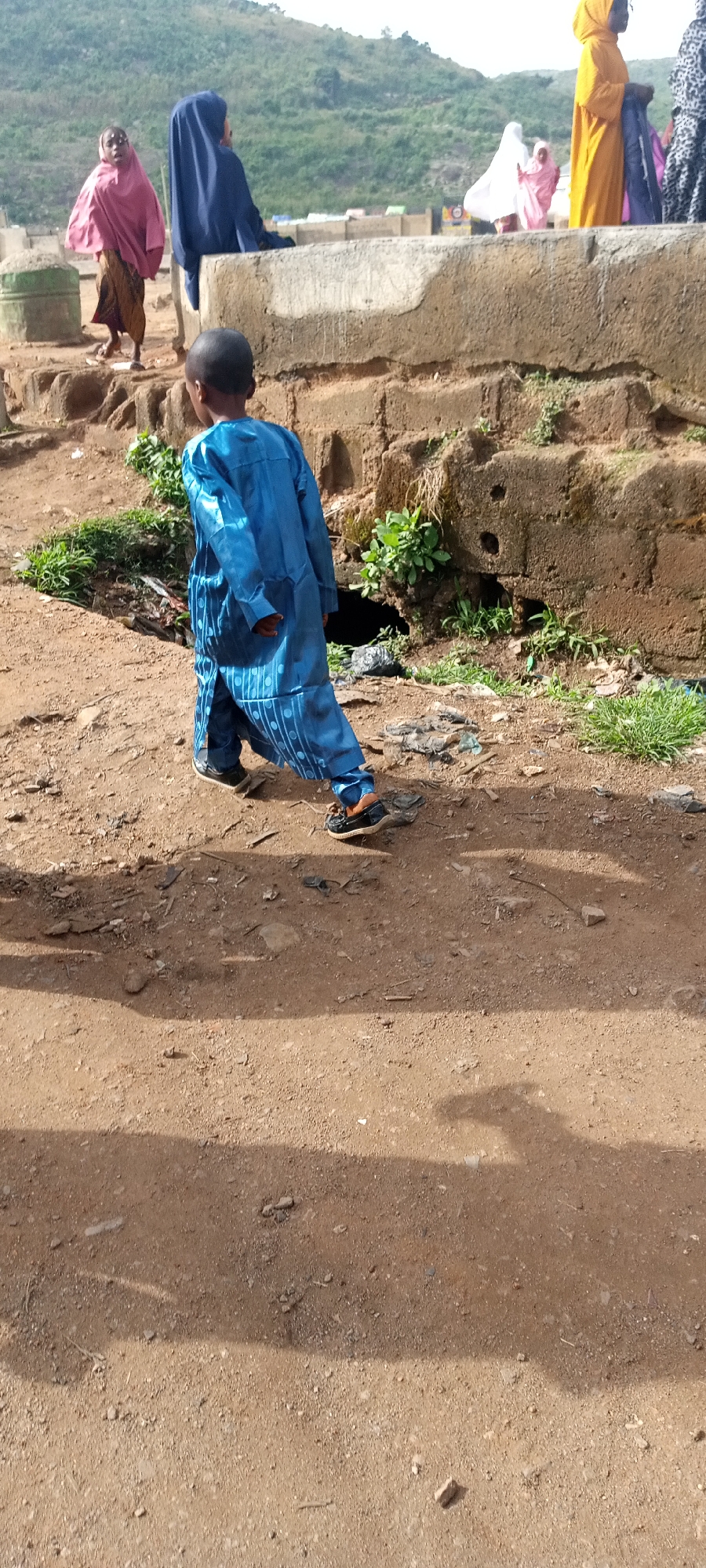 The image shows a child wearing a blue traditional outfit walking away from the camera on a dirt road in Naraguta Hausawa, Nigeria. The child is near a concrete structure. In the background, there are several people wearing traditional garments, and a hill covered in vegetation.