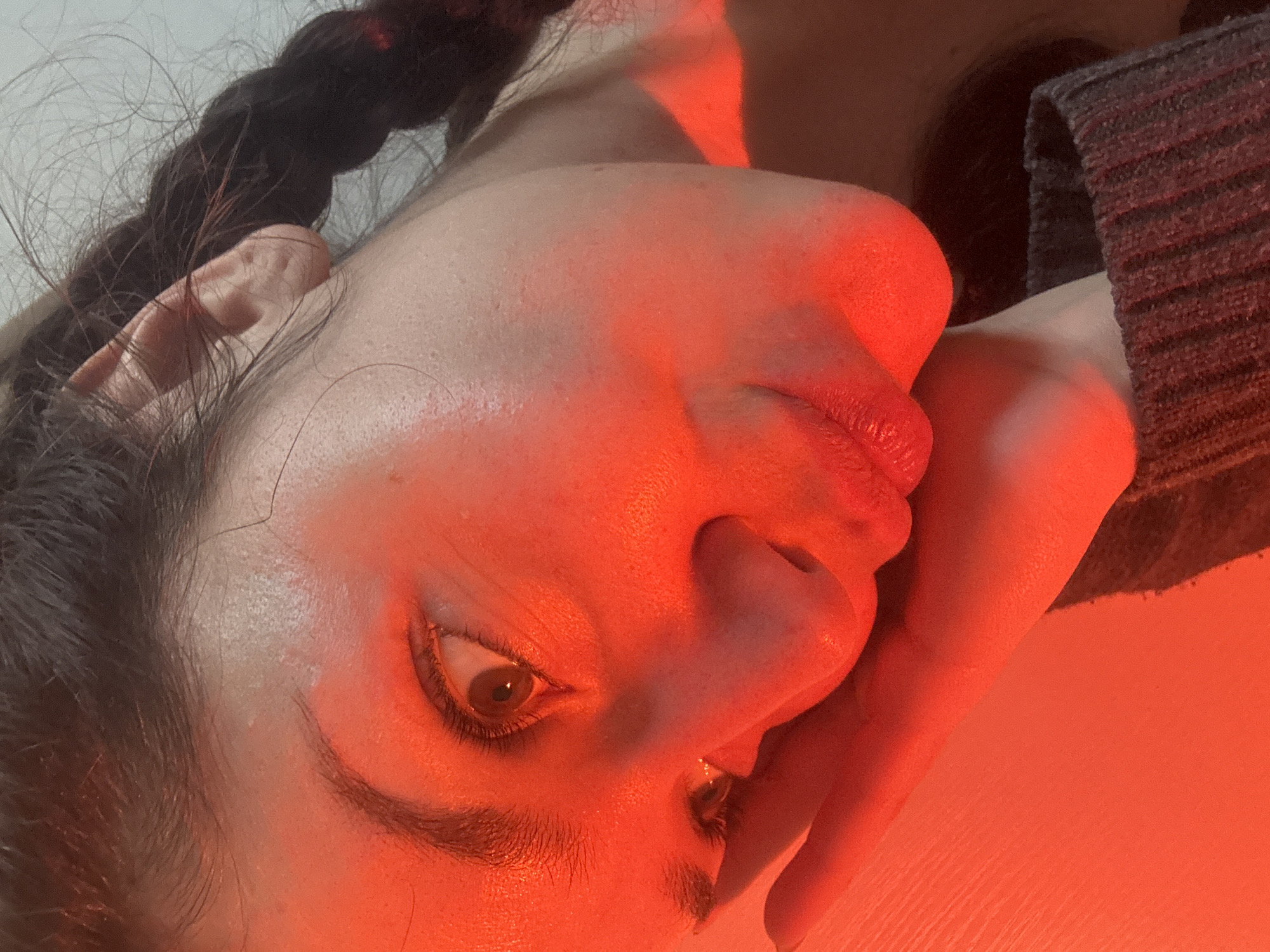 A close-up, tilted portrait of a woman with brown hair braided and resting to the left. Her face is illuminated by red lighting. Her right eye is visible and focused. Her hand is raised to her face. She is wearing a gray sweater. The background is a plain white surface. The location is not discernible from the image.
