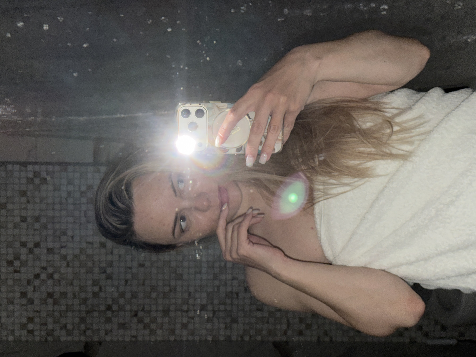 An image, rotated 90 degrees counter-clockwise, depicts a woman taking a mirror selfie with a smartphone. The woman, who has blonde hair and light skin, is wrapped in a white towel, suggesting she is in a bathroom or has recently bathed. Her left hand holds the smartphone, which has a multi-lens camera system, a pop socket or stand on its back, and a decorative case. The phone's flash is prominently activated, creating a bright white glare and lens flares across the image. Her nails are manicured with a light, possibly white or clear, polish. Her right hand is positioned near her chin.
The background, visible in the mirror's reflection, consists of small, dark-toned square tiles, typical of a bathroom wall. The mirror itself appears to have specks or dust on its surface. The lighting is primarily from the phone's flash, illuminating the woman and the immediate reflection. The woman's expression is neutral as she looks towards the camera. No discernible text is visible on any object. The overall scene conveys a private, intimate moment of self-capture.