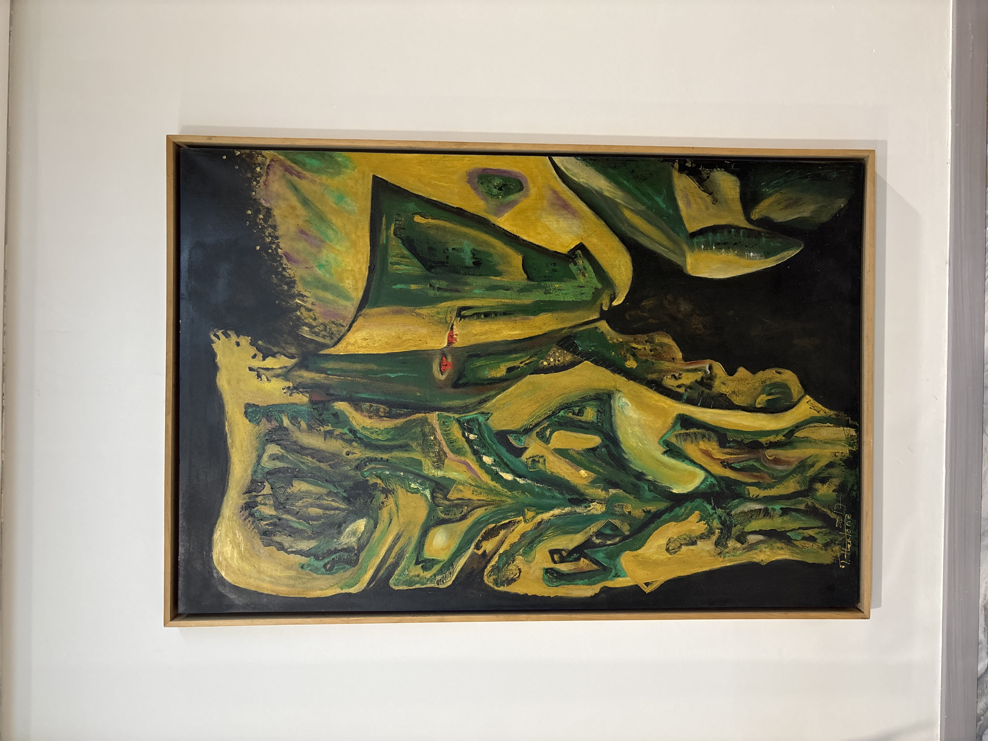 The image shows a framed abstract painting with predominantly yellow and green hues, set against a black background. The painting features organic, flowing shapes and appears to have a textured surface. The artwork is hung on a white wall, and a light gray baseboard is visible at the bottom of the frame. In the lower right corner of the painting, a signature and date are faintly visible: "T. Hardjo 2017". This artwork is displayed in the city of Binong, Indonesia.