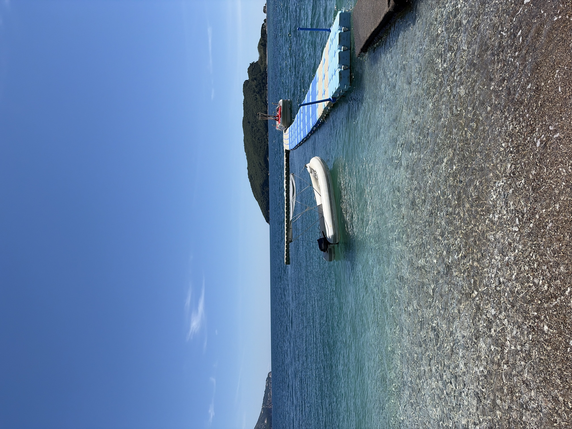 The image depicts a coastal scene in Budva, Montenegro. A clear blue sky dominates the upper left portion of the frame. The Adriatic Sea occupies the central and right portions of the image, with varying shades of blue and turquoise suggesting depth. Two small boats, one a white inflatable dinghy with an outboard motor, are visible. They are moored near a blue and yellow plastic pontoon extending from a dark concrete quay or pier. The shoreline consists of a pebble beach that transitions into the water. An island covered with green vegetation is situated in the background, slightly obscured by the haze. There are no people visible in the image. The overall impression is of a tranquil, sunny day at the coast.