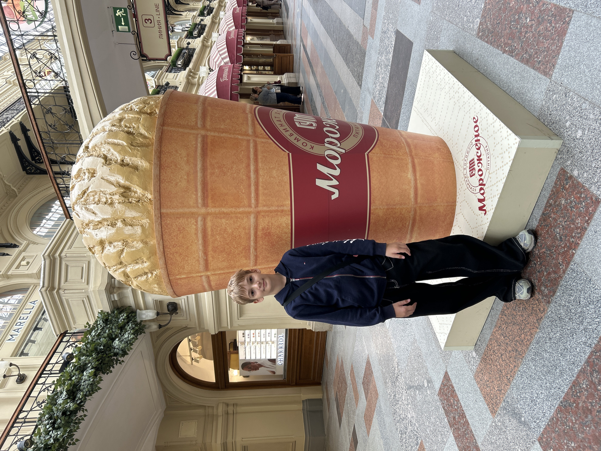 The image captures a boy posing in front of a giant ice cream cone sculpture inside the GUM department store in Moscow, Russia. The GUM, located on Red Square, is an iconic historical shopping mall with a grand, arched glass roof and intricate architecture. The ice cream cone, which appears to be a replica of a classic ice cream, is a prominent display. The boy, wearing a dark blue hoodie and black pants, stands on a light-colored pedestal that supports the sculpture. The floor is a mosaic of polished stone tiles in various shades of grey, brown, and red. In the background, there are store facades, balconies with greenery, and illuminated signage. The overall scene suggests a casual moment captured during a visit to a popular tourist attraction.