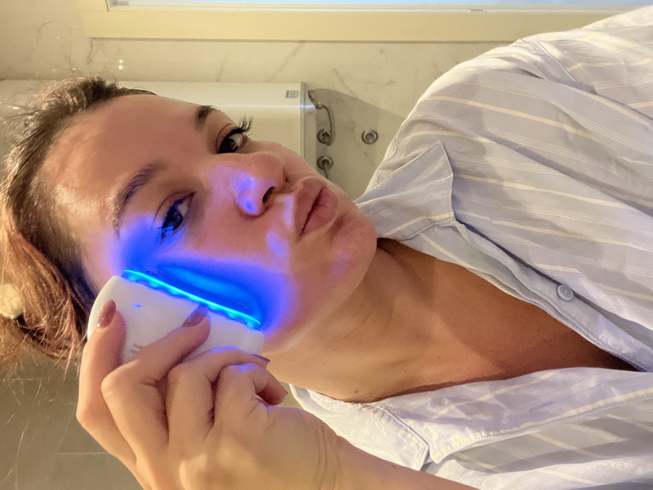 A female subject is depicted from the chest up in an indoor tiled room, identified as a bathroom within the city of Budva, Montenegro. The subject has light brown hair and dark eyes, and is wearing a light blue and white striped button-down shirt. Her right hand, displaying manicured nails with light polish, holds a white, ergonomic electronic device against her left cheek and jawline. The device emits a bright blue light that illuminates the surrounding skin. The subject is looking directly at the camera with her lips slightly puckered. The background features white marble-patterned wall tiles, a white wall-mounted water heater or boiler to the right, and two circular wall fixtures beneath it.