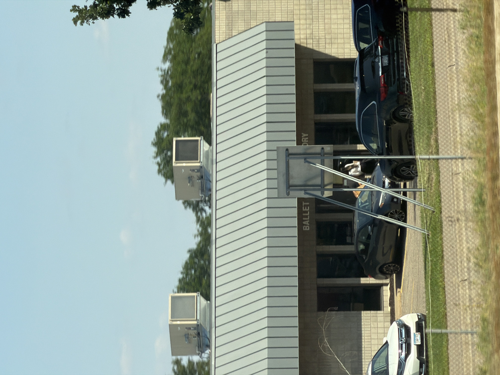 The image shows the exterior of a building with a light-colored brick facade and a corrugated metal awning. Above the awning are two large, beige HVAC units. The word "BALLET" is visible in large letters above a set of windows on the building. In front of the building, several cars are parked in a lot. A white SUV is parked on the left, with a dark gray sedan to its right. Further to the right, a black SUV is parked, and another dark-colored vehicle is partially visible behind it. A chain-link fence runs along the bottom of the image, with a grassy area and some dead branches in front of it. The sky is a clear blue with a few wispy clouds. The scene appears to be a commercial or institutional building, possibly a dance studio or performing arts center, located in Saint Paul, United States of America.