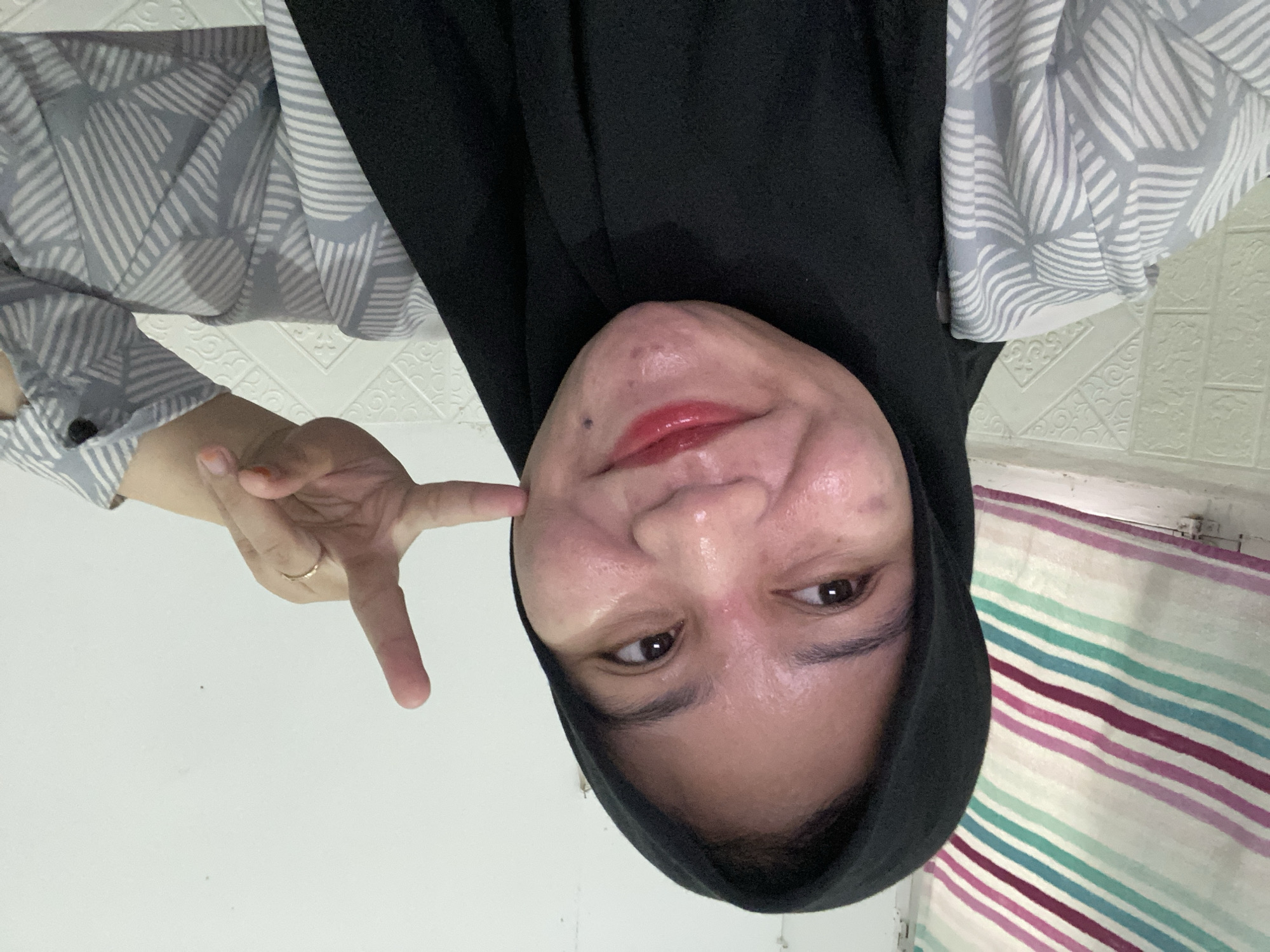 A close-up selfie shows a young Indonesian woman, likely in her early twenties, wearing a black hijab and a light grey and white patterned shirt. She has brown eyes and is smiling, with red lipstick on her lips. Her right hand is raised, making a peace sign near her cheek. The background features a wall with a subtle textured pattern and what appears to be a striped curtain or fabric on the left side of the frame. The lighting suggests an indoor setting, possibly a home environment. The location is Banjarbaru, Indonesia.