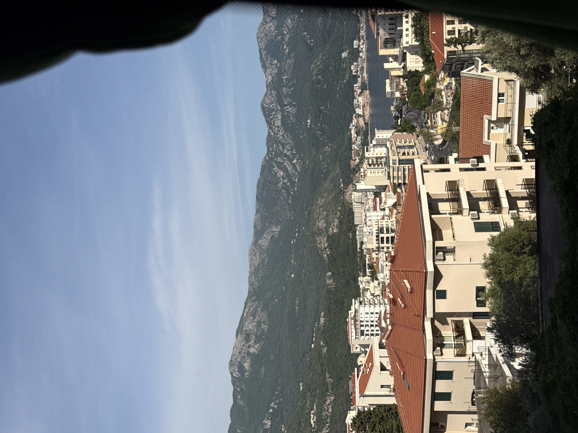 The image shows a view of Boreti, Montenegro, from an elevated position. The scene includes mountains, a body of water, and the town's architecture. The mountain range, densely covered in vegetation, runs parallel to the shoreline and the town. Buildings, mostly residential, with light-colored facades and red-tiled roofs, are clustered along the coast. A waterway, possibly a river or a bay, runs through the town. The sky is clear and blue. The view is partially obstructed by dark, undefined shapes at the top of the frame, likely part of an interior.
