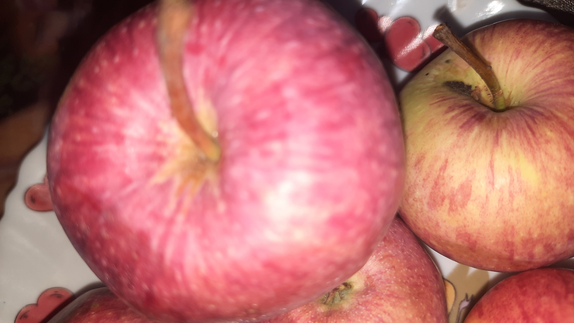 Four apples are visible in the image, arranged in a pile on a white plate with a heart-shaped pattern. The apples are primarily red and yellow, with some streaking. One apple in the foreground is a solid red and takes up a significant portion of the frame. To its right is another apple that is yellow with red streaks. Two other apples are partially visible at the bottom of the frame. The lighting suggests an indoor setting, possibly a kitchen. There is no visible indication of the city of Шклоў, Belarus.