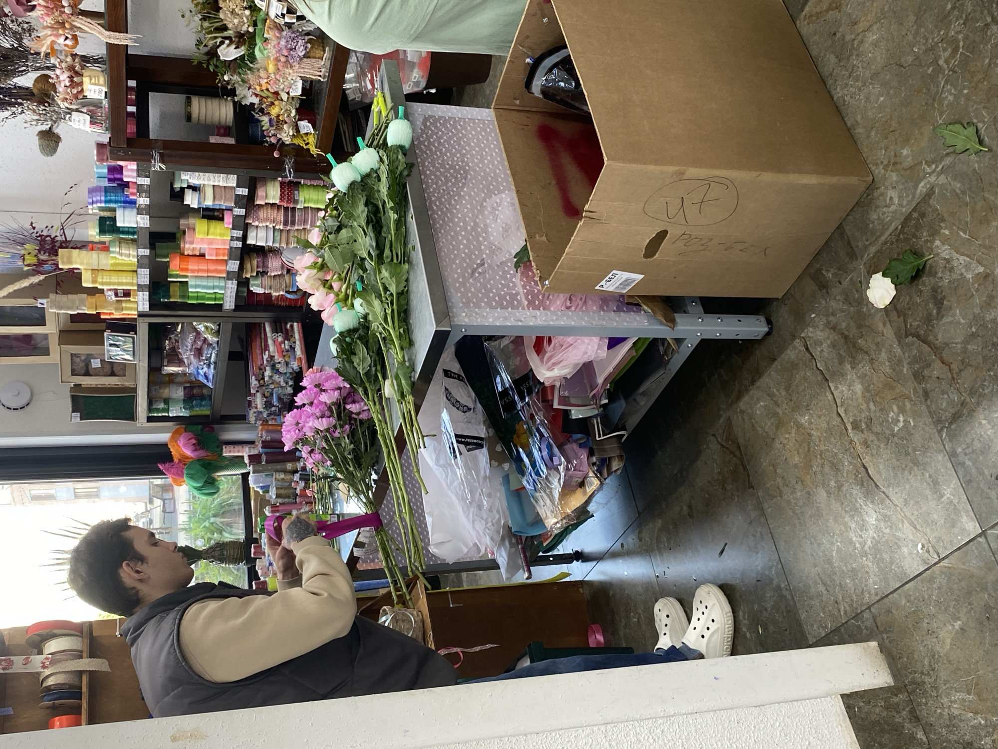 A man is seen wrapping pink ribbon around flower stems in a florist shop. The man wears white Crocs and a vest and sits behind a white counter. Several different colored ribbons are on a shelving unit behind the counter. A tall gray metal shelf holds a variety of flower bouquets, vases, and supplies. A large cardboard box is on the right of the shelf. The floor is dark, mottled, brown tile.