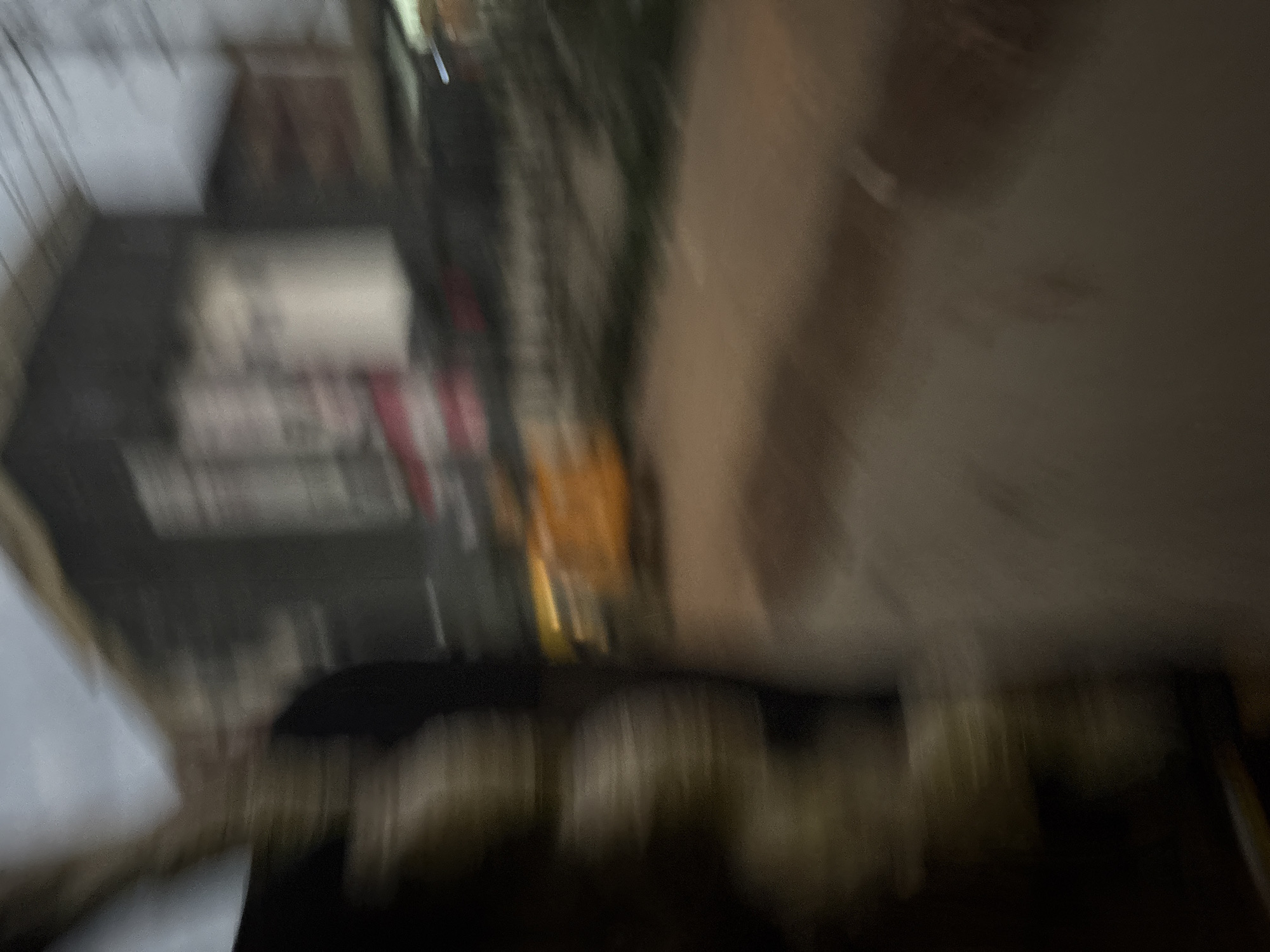 The image depicts a blurry, motion-induced scene in Onitsha, Nigeria. In the foreground, a person or object with a textured, stacked appearance is visible on the left, appearing dark and somewhat cylindrical. To the right of this, a bright orange object, possibly a vehicle or a part of one, is blurred and elongated horizontally. The background consists of indistinct buildings with dark facades and lighter rectangular shapes that could be windows or signs. A power line is faintly visible overhead. The overall impression is of movement, possibly captured from a moving vehicle at dusk or night. The ground appears to be a dirt or paved surface, with a reddish-brown hue. The lighting is dim, contributing to the motion blur and lack of clear detail.