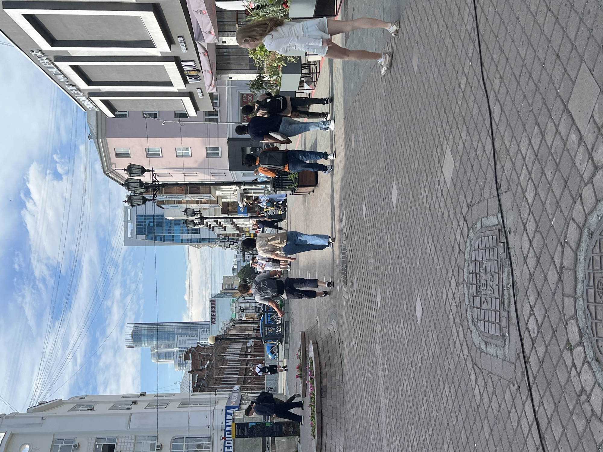 The image shows a street scene in Yekaterinburg, Russia, on a sunny day. The perspective is from above, looking down at the street and the people walking on it. The street is paved with grey diamond-shaped tiles, and there are some manhole covers visible. On the left side of the street are buildings of various architectural styles, some modern and some older. Power lines are visible crisscrossing the sky.
Several people are walking along the street. In the foreground, a person in dark clothing is walking away from the camera. Further down the street, a group of people are walking in the same direction. One woman is wearing a white dress and sandals. Another person is wearing a dark t-shirt and jeans, carrying a backpack. There are other individuals and groups scattered along the street, some appearing to be talking or looking at their phones.
The overall atmosphere seems to be one of a normal, bustling day in a city. The lighting suggests it is daytime, likely late morning or early afternoon, given the brightness and the shadows cast by the buildings and people. There is no specific event or organized activity apparent; it appears to be people going about their daily routines. Some signage is visible on the buildings, but it is difficult to read clearly due to the resolution and orientation. One sign on a building in the distance appears to have Russian text.