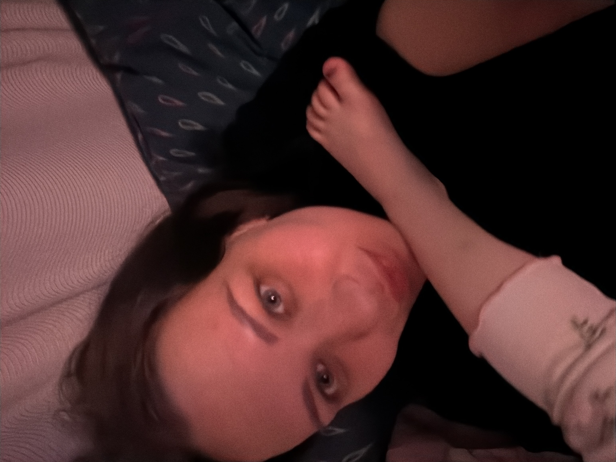 The image shows a close-up of a woman lying down, with a child's foot resting on her arm. The woman's face is visible, with her eyes looking towards the camera. The lighting is dim and casts a reddish hue over the scene. The background appears to be a bed with patterned bedding. There are no specific landmarks or contextual clues that would indicate the location in the city of Perm, Russia. The image is a candid shot, likely taken indoors in a residential setting.
