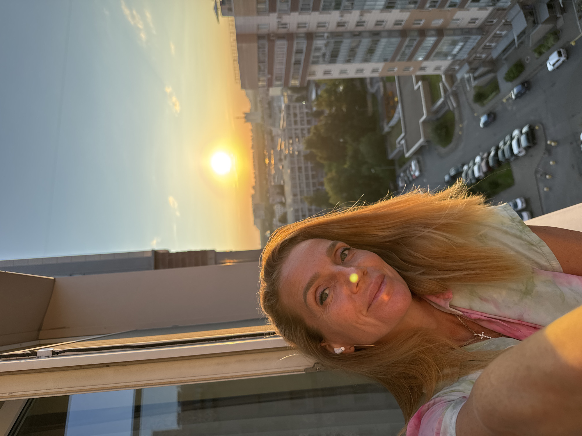 A woman with blonde hair is taking a selfie on a balcony at sunset. She is wearing a tie-dye shirt and a cross necklace. In the background, the sun is setting over a city skyline with apartment buildings and trees. The scene appears to be in a residential area in Russia.