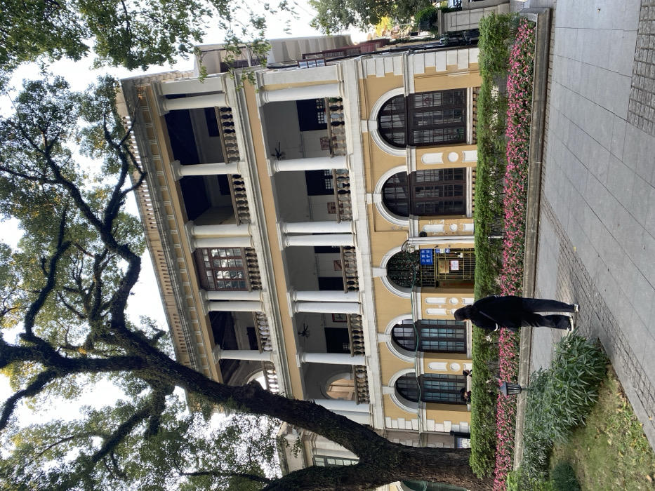 A large, multi-story building with a distinctive architectural style dominates the scene, partially obscured by a mature tree in the foreground. The building features a light yellow facade adorned with prominent white columns supporting multiple levels of open balconies or verandas, some with white balustrades. The ground floor displays large, dark-framed arched windows, while the upper levels have rectangular windows and visible ceiling fans within the covered balcony spaces.
On the left, a large, old tree with a thick, gnarled trunk and extensive branches covered in moss or lichen fills a significant portion of the frame, its green foliage silhouetted against a bright, clear sky.
In front of the building, a well-maintained garden bed featuring green shrubs and small pink flowers lines a paved sidewalk. A person, dressed in dark clothing and possibly carrying a backpack, stands on this sidewalk, facing away from the camera and looking towards the building.
The image is well-lit, indicating daytime. A blue sign visible on the building's entrance clearly displays Chinese characters "出入口" (chū rù kǒu), which translates to "entrance/exit." The setting suggests an urban environment, consistent with the specified location of Guang Zhou Shi, China, showcasing a blend of natural elements and historical architecture.
