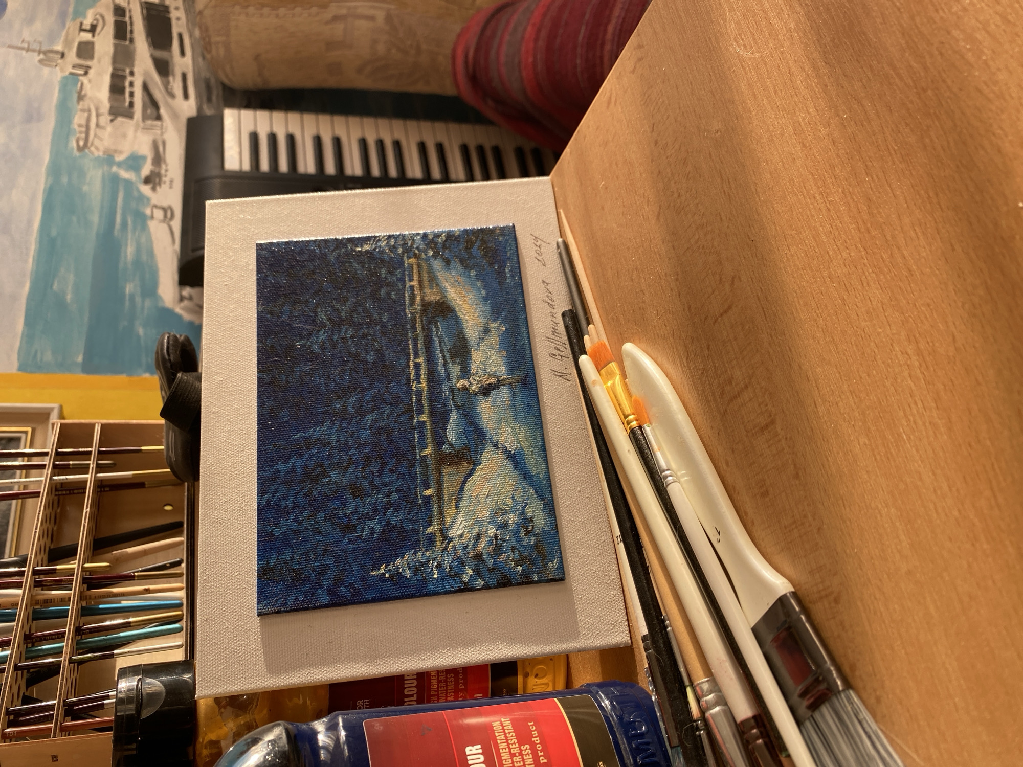 The image depicts an artist's workspace, likely in Bílina, Czechia, with a focus on a small, newly completed painting. The painting itself is a winter scene, featuring a snow-covered bridge over a stream, surrounded by dark, snow-laden trees, and a lone figure walking on a path. The artwork is signed "M. Gellmundova 2024".
The painting is placed on a larger, light-colored canvas, leaning against a wooden surface. In the foreground, a collection of paintbrushes are arranged neatly, some with bristles showing signs of use. To the left, a dark blue bottle of "COLOUR" art pigment is visible, with labels indicating it is a highly pigmented, water-resistant product. Behind the painting, a blurred backdrop includes a partial view of a yacht painting, a keyboard, and a glimpse of a person's leg covered in a red and white striped blanket. The overall lighting suggests a warm, indoor environment, possibly illuminated by natural light from a window, casting shadows across the wooden table. A wooden organizer filled with more art supplies is visible in the upper left portion of the image.