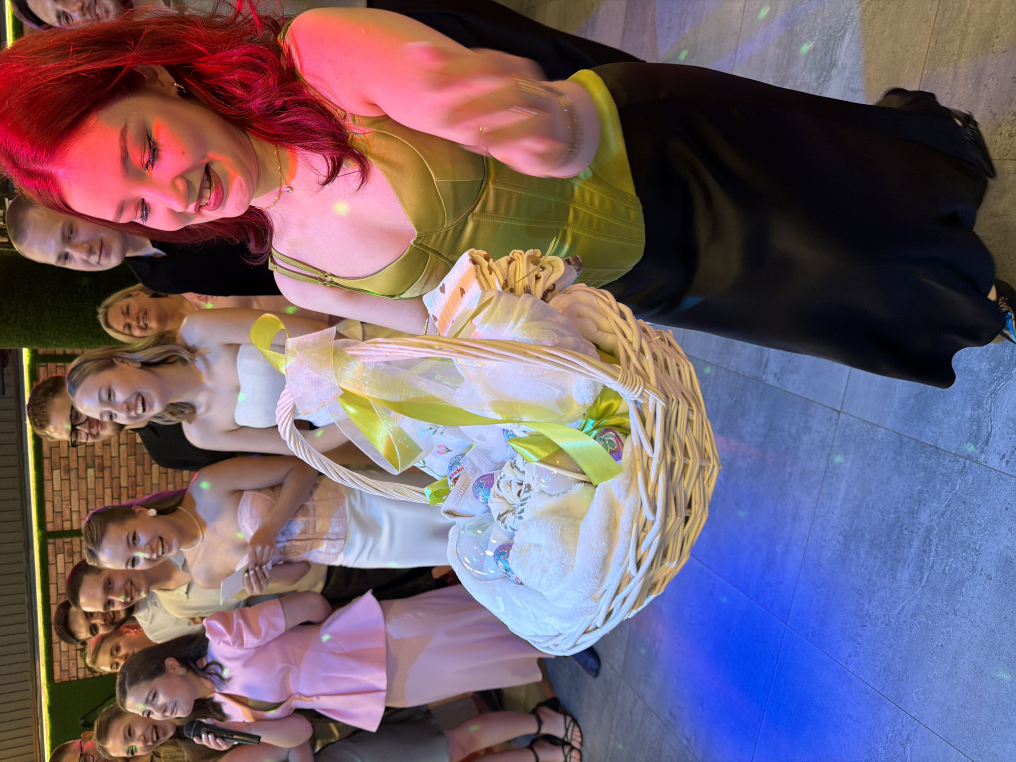 In Chernushka, Russia, a group of young adults are gathered indoors, likely at a celebration or party. The foreground is dominated by a young woman with vibrant red hair, illuminated by pink and red lighting, wearing a lime green corset-style top and a long black skirt. She is smiling and holding a wicker basket filled with gifts, including what appear to be baby items, wrapped in white fabric and adorned with yellow and green ribbons. Behind her, several other individuals are visible, including a woman in a pink outfit, a woman in a strapless white top, and various men and women in casual attire. The background features a brick wall and a dense green hedge. The floor is tiled and reflects the blue and green lighting.