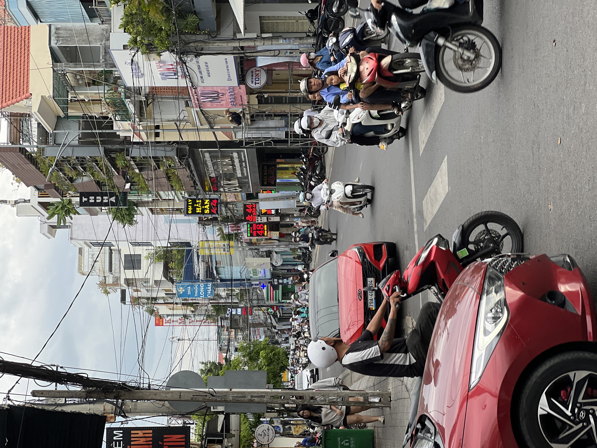 This is a street-level, eye-level medium shot of a busy urban scene in Nha Trang, Vietnam, likely on a cloudy day. The foreground is dominated by the front of a red car, showing its headlight and grille. To the right of the car, a motorcycle with a rider wearing a white helmet is visible, and further right, another motorcycle with a rider wearing a dark helmet and a black and white shirt is also present. In the mid-ground, a red car is stopped at a crosswalk. Numerous people are on motorcycles, some wearing helmets and masks, navigating the busy street.
The background shows a dense collection of multi-story buildings lining both sides of the street. These buildings have a mix of architectural styles, with some featuring balconies and greenery, and others appearing more commercial with signs. The sky is overcast, and a complex network of electrical wires crisscrosses overhead, a common feature in urban Vietnam. Signs in Vietnamese are visible on the buildings, indicating shops and businesses. The overall impression is one of a vibrant, active city street.