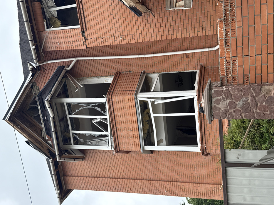 A red brick building is damaged, particularly around the windows. Two large windows on the second story are missing glass, with cracked frames and debris visible inside. Brickwork shows signs of cracking. Part of the roofline above the windows is damaged and missing pieces. A white pipe runs along the exterior wall, and a fence is visible in the foreground. The building appears to be facing outward, as if seen from a low angle perspective.