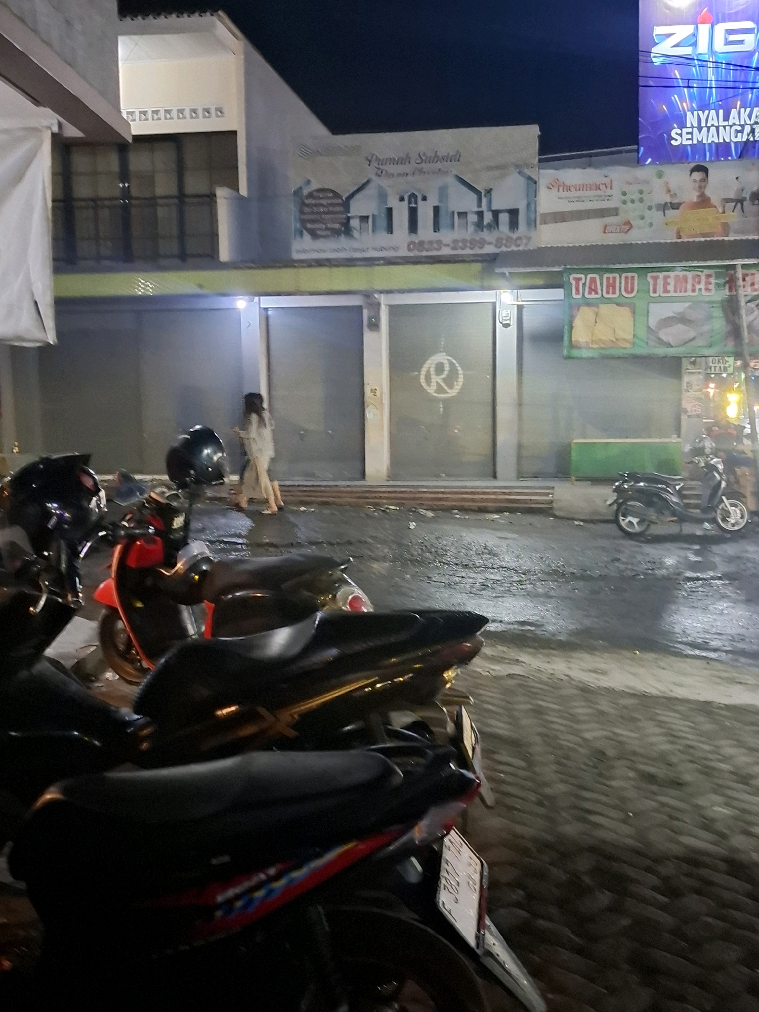 The image shows a nighttime street scene in Ciheulang Tonggoh, Indonesia, with several motorcycles parked in the foreground. In the middle ground, two women are walking past a building with large roll-up doors. Above the doors, a banner advertises "TAHU TEMPE," indicating a food stall or eatery. Other signs are visible, including one that reads "Rumah Subsidi" and another with "Rheumaviv." In the background, a neon sign for "ZIG" is illuminated. The street is wet, suggesting recent rain. The overall atmosphere is one of a typical Indonesian street at night, with commercial activity and parked vehicles.