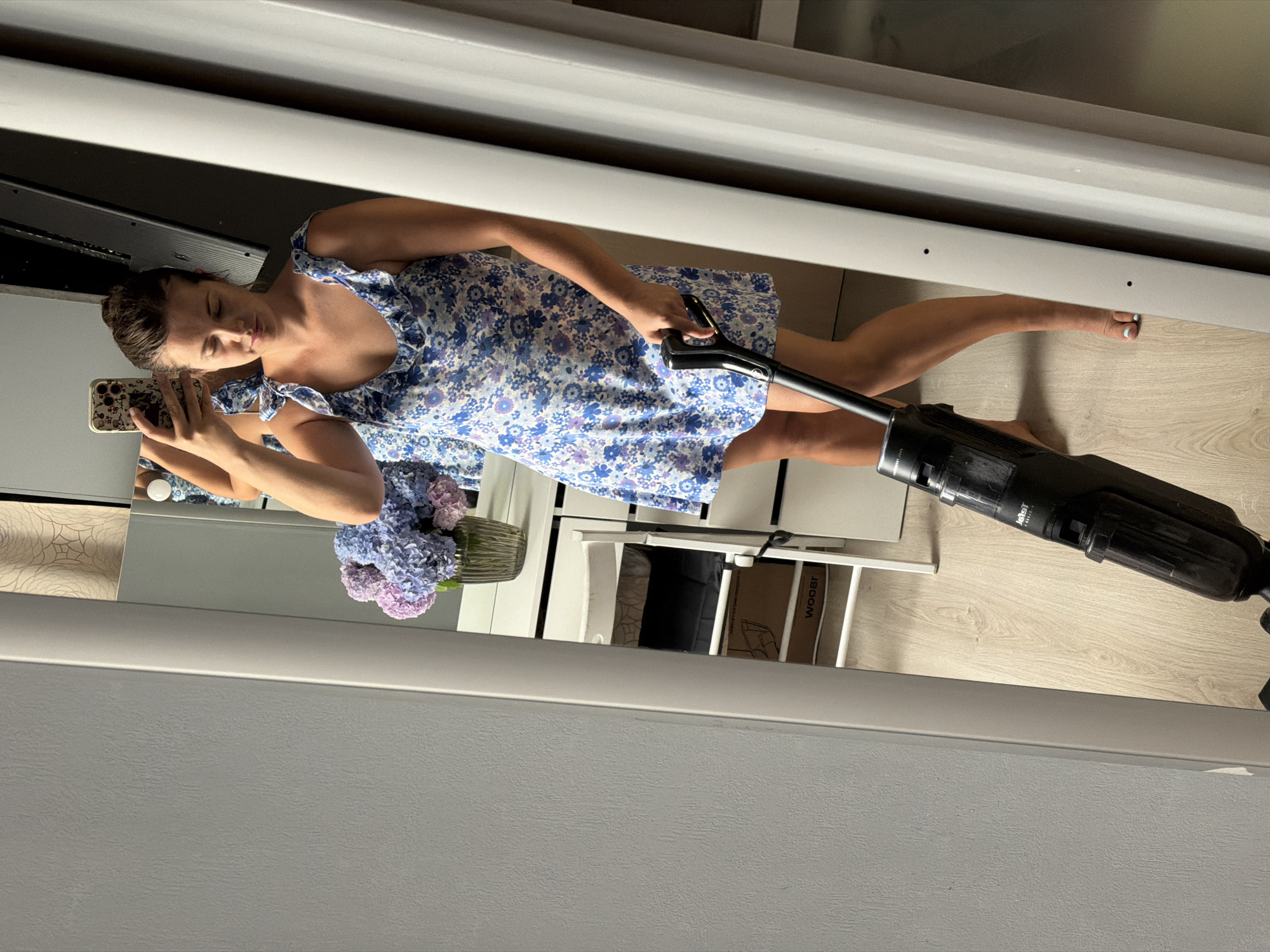 A woman wearing a blue floral dress is vacuuming her floor. She is holding a black vacuum cleaner and looking at her reflection in a large mirror. Behind her, a vase of hydrangeas sits on a dresser. The scene appears to be indoors, possibly in a bedroom or living area. The floor is made of light-colored wood.
