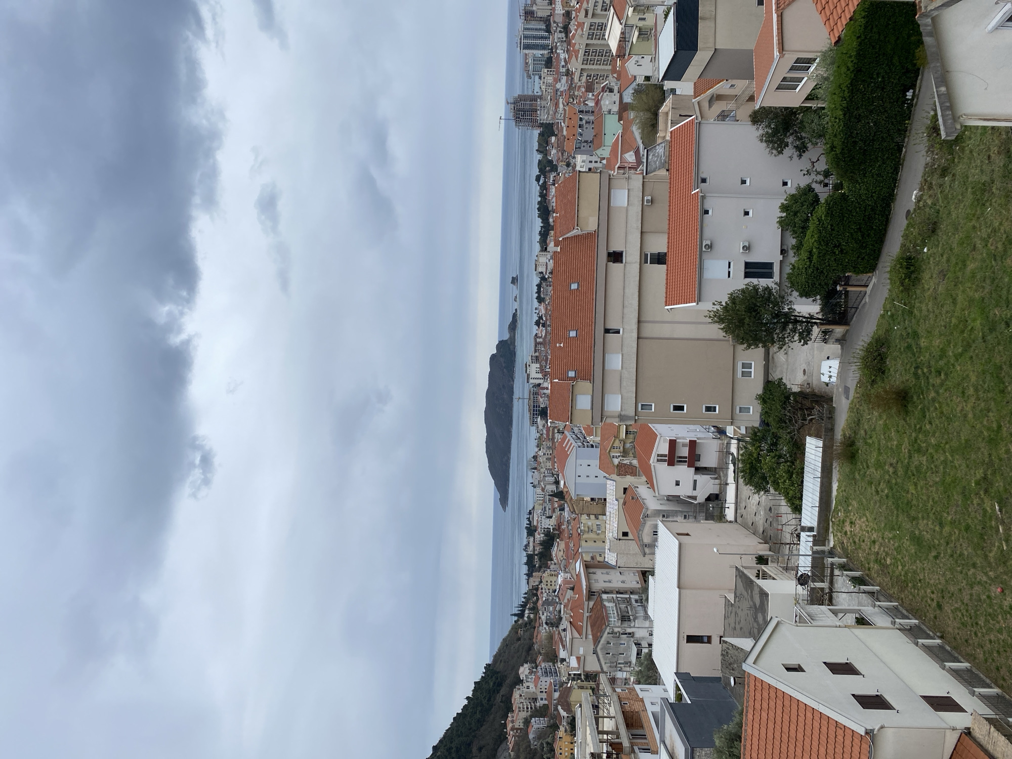 The image depicts an elevated view of Budva, Montenegro, from an angle looking towards the sea. The scene is dominated by overcast skies. The Adriatic Sea is visible, with Sveti Nikola Island in the distance. The foreground features buildings with red-tiled roofs characteristic of the Mediterranean architectural style. Green patches of vegetation dot the landscape, and a small grassy area is present on the right side of the frame.