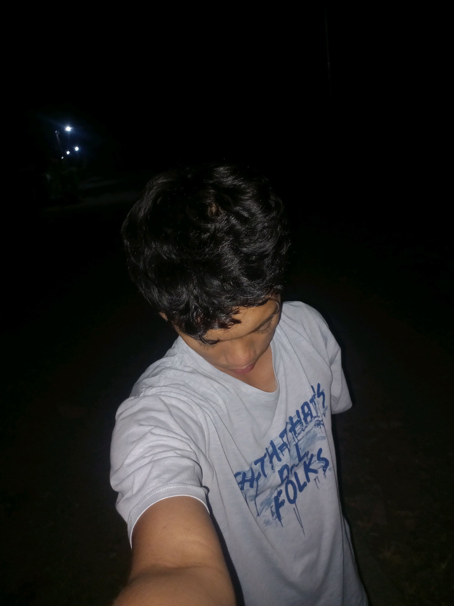 The image shows a young person from the chest up, looking down. They are wearing a light-colored t-shirt with blue text that reads "THAT'S ALL FOLKS". Their hair is dark and wavy. The background is dark, with a few distant lights visible, suggesting it is nighttime. The location is identified as Gambiranom, Indonesia.