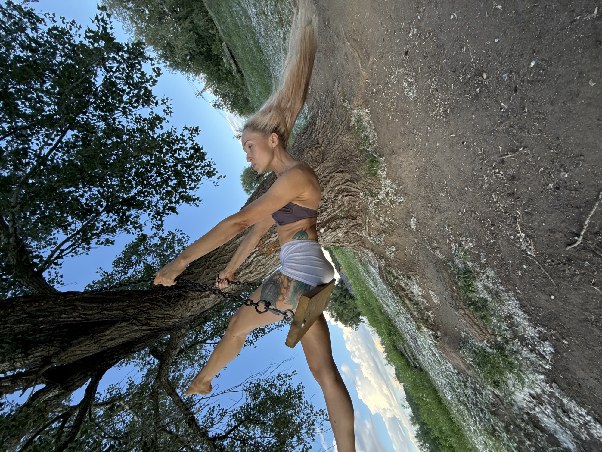A blonde woman is swinging on a wooden swing suspended from a large tree. She is wearing a dark bra and light shorts, and her legs are outstretched in a split. She has a tattoo on her thigh. The swing is attached to a thick chain. The tree is large with textured bark and is surrounded by grass and dirt. There are some white fluffy materials scattered on the ground around the tree. The sky is blue with some clouds. The scene appears to be outdoors, possibly in a park or natural area. This location is not identifiable as Chaikovsky, Russia based on the provided image.