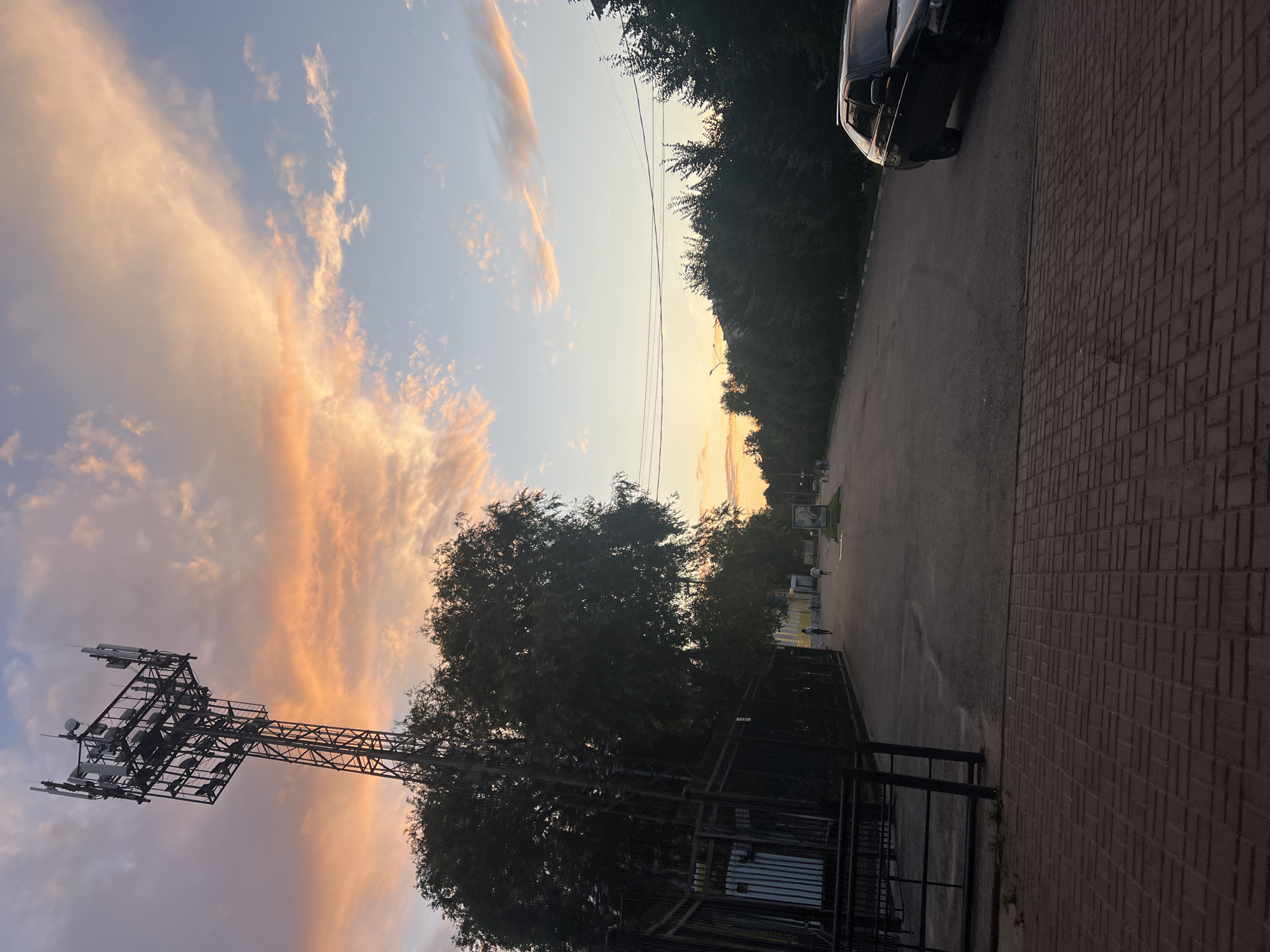 An outdoor, ground-level, medium shot captures a road and sidewalk in Ulyanovsk, Russia. The sky displays a sunset with hues of pink and orange clouds against a pale blue backdrop. A large telecommunications tower stands on the left side of the image, topped with antennas and equipment, adjacent to a fence and a large tree. The road, paved with asphalt, extends into the distance, lined by trees on the right. A black station wagon is parked on the right side of the road. Several indistinct figures are walking on the sidewalk in the middle ground. The foreground is dominated by a brick-patterned sidewalk.