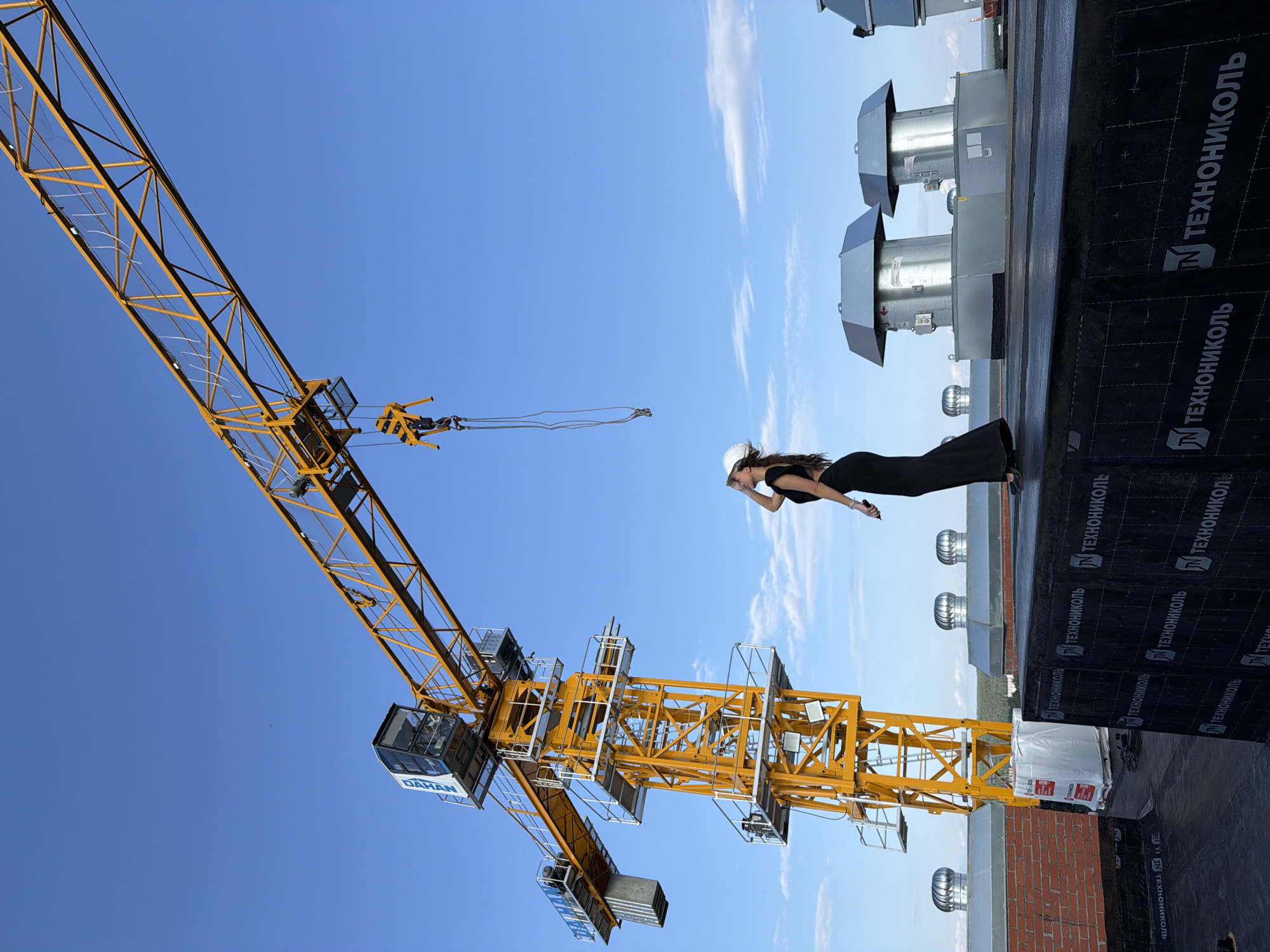 A woman wearing a black dress and a white hard hat stands on a rooftop, leaning forward with her arm extended. She appears to be looking down at something below. Behind her, a large yellow construction crane dominates the scene, its boom stretching across a clear blue sky with scattered white clouds. The rooftop is covered with black material, likely roofing, and features several large ventilation units. The woman is positioned in the foreground, with the crane and the rooftop elements behind her. The image seems to be taken during the day, with bright sunlight casting shadows. The word "ТЕХНОНИКОЛЬ" is visible in white text on the black roofing material, repeated multiple times. The brand "DAHAN" is also visible on the operator's cabin of the crane. The scene suggests an active construction site or a completed building with ongoing work. The woman's pose and attire, combined with the construction equipment, create a striking contrast.