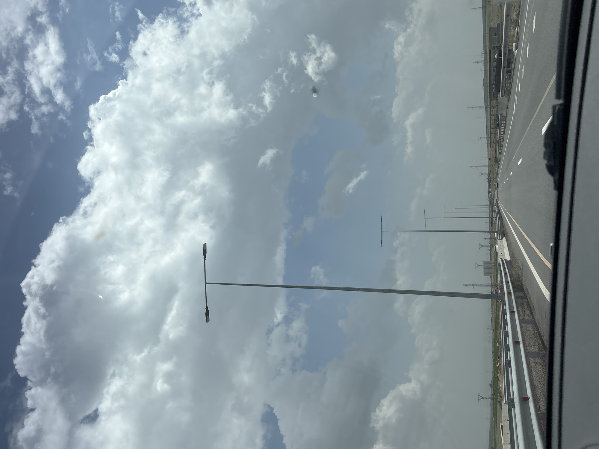 The image shows a highway in the Talin region of Armenia, viewed from inside a moving vehicle. The sky is filled with predominantly gray and white clouds with patches of blue. A straight asphalt road with clear lane markings stretches into the distance, flanked by metal guardrails and grassy areas. Streetlights are evenly spaced along the road. Power transmission towers are visible further away in the distance. The focus is sharpest on the immediate road surface and the nearby guardrails. A portion of the vehicle's interior is visible on the right edge of the frame.