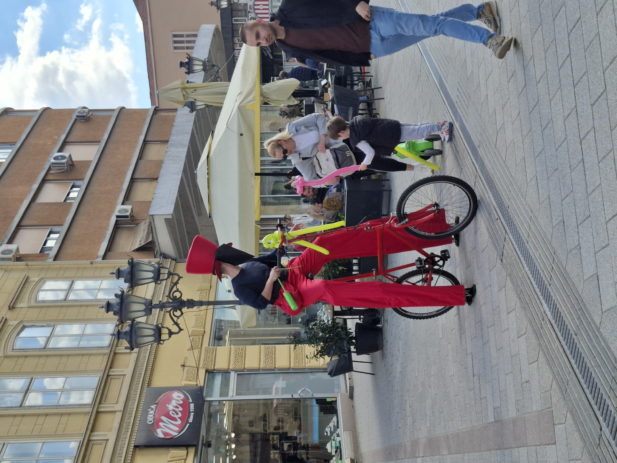 An outdoor street scene in Novi Sad, Serbia shows a performer wearing a red suit and tall hat, standing next to a red bicycle with balloon animals attached. The performer appears to be interacting with a woman and child who are holding balloons. Behind them are tables with umbrellas and people sitting. In the background is a man walking, and buildings with windows and air conditioning units. A street light and a storefront with the name "Metro" in Cyrillic are also visible. The ground is paved with grey bricks.