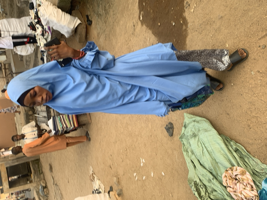 The image depicts an outdoor market scene in Okene, Nigeria, characterized by an unpaved, dusty reddish-brown ground surface. In the foreground, a woman walks from right to left, captured mid-stride. She is wearing a light blue hijab covering her head and body, with a black underscarf visible at the forehead. Her left arm is slightly bent, holding a black smartphone vertically in her hand, with red beaded bracelets visible on her wrist. She wears black sandals and patterned black and white trousers visible beneath her blue garment.
In the mid-ground, to the left of the walking woman, a stack of green fabric lies on the ground, with a smaller bundle of light-colored patterned fabric on top. Further into the mid-ground, on the left, two men are visible. One man, facing the camera's left, wears an orange long-sleeved tunic and matching trousers. The other man, slightly behind him, wears a light-colored shirt and trousers, standing behind a wooden stall displaying neatly folded stacks of multi-colored fabrics.
In the background, on the right side, there is a raised area with several large white sacks and various items, including shoes and clothing, scattered on the ground. A dark, possibly black, object resembling a firearm or tool is partially visible near the base of this raised area. Further back, architectural elements like steps and building structures are discernible, with other blurred figures moving in the distance. The overall scene suggests a busy, open-air retail environment during daylight hours.