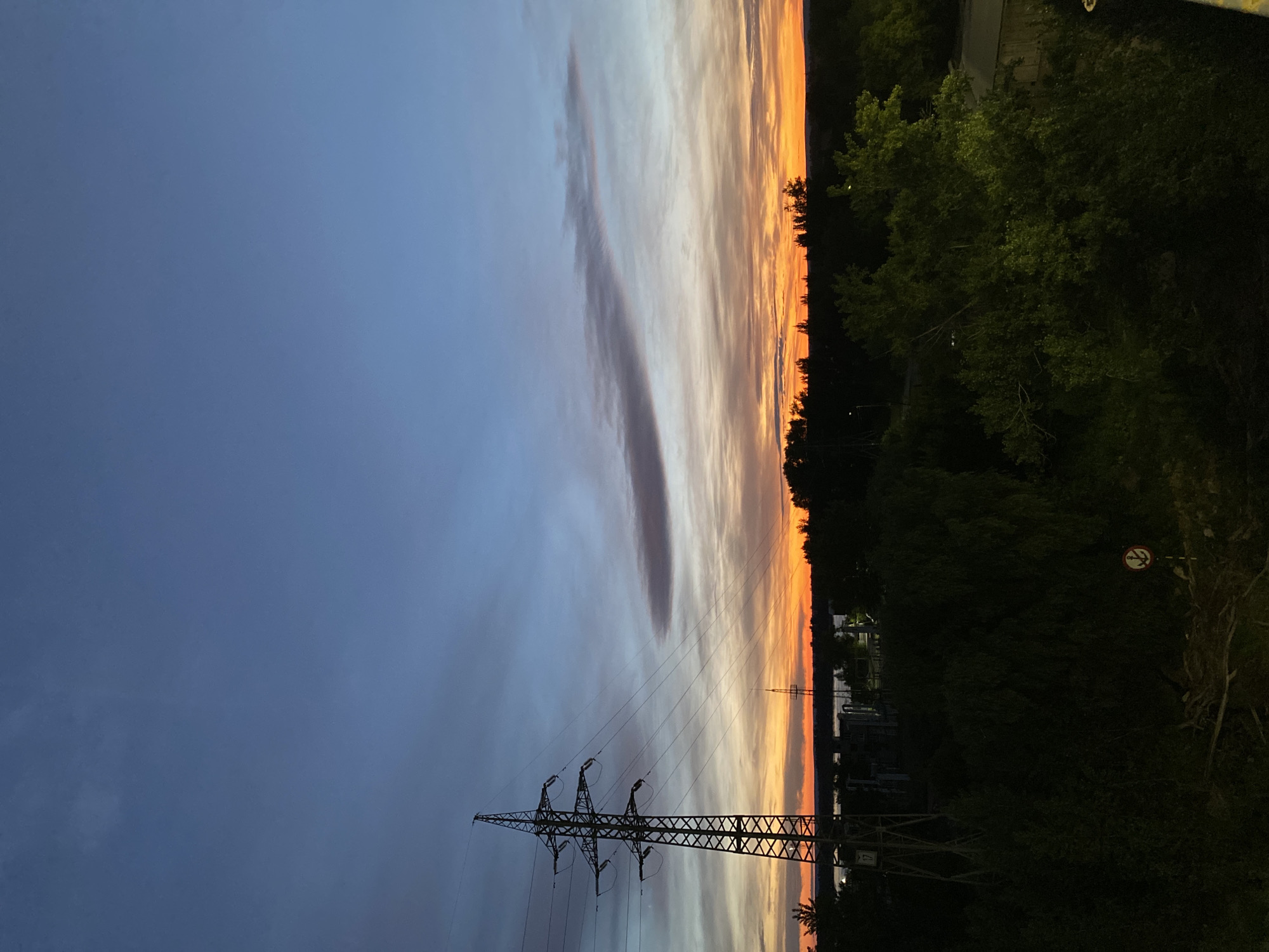 This is a vertical, high-angle shot of a sunset over Novokuybyshevsk, Russia. The sky is filled with a dramatic gradient of colors, from deep blue on the left to vibrant orange and yellow on the right, indicating sunset or twilight. Wispy clouds are scattered across the sky, catching the last rays of the sun. In the foreground, a large, lattice-style electricity pylon stands prominently, silhouetted against the colorful sky. The pylon is supported by wires that extend towards the horizon.
To the right of the pylon, a dense line of dark green trees forms the edge of the landscape, with some lighter green foliage visible. Beyond the trees, faint structures and a distant pylon can be seen. The overall scene evokes a sense of calm and the beauty of nature at dusk, juxtaposed with the industrial element of the electricity pylon. The lighting suggests it is either early morning or late evening. No people are visible in the image. There is a small circular sign with a prohibition symbol on the right side of the image.