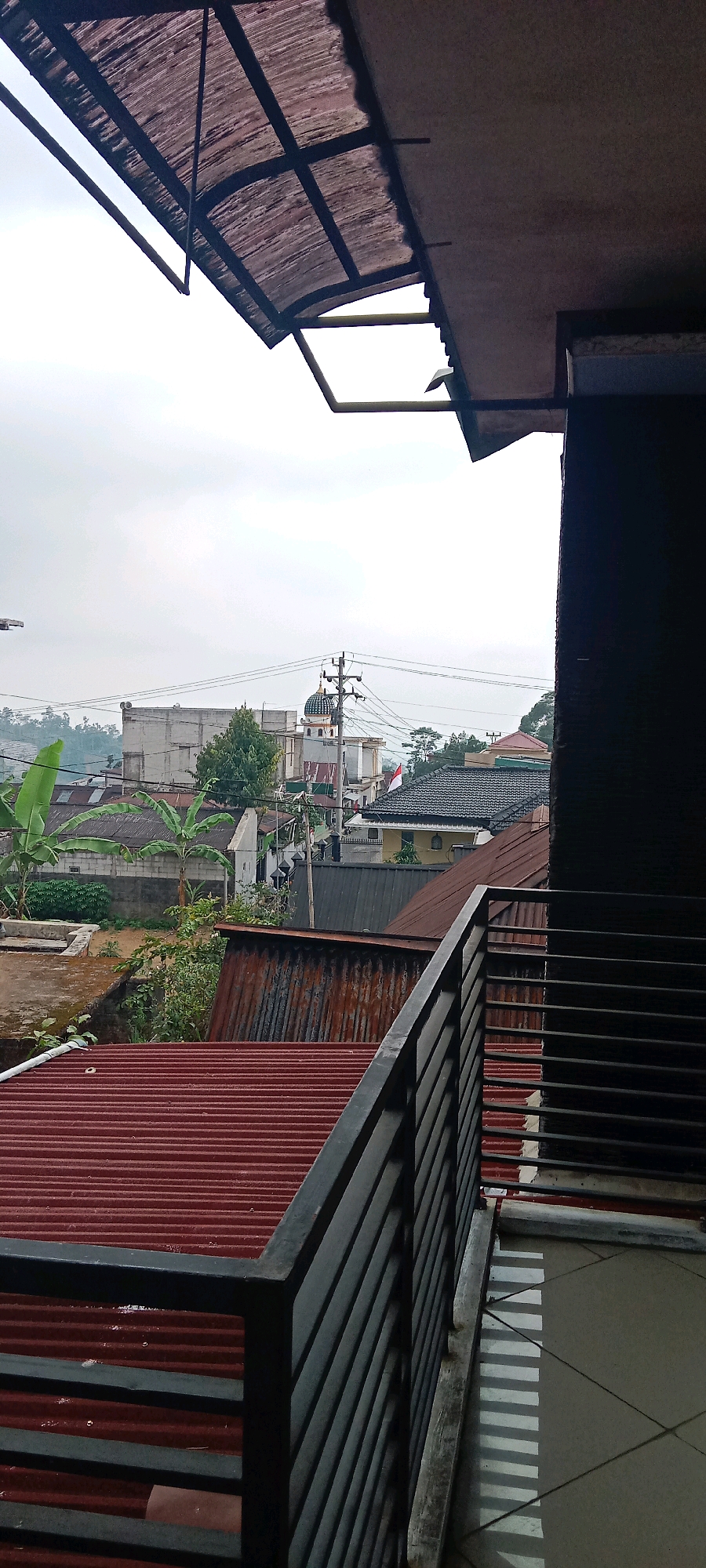 The image is a low-angle, full shot taken from a balcony overlooking a residential area in Krinjing, Indonesia. The foreground is dominated by the dark metal railings of the balcony and the reddish-brown corrugated metal roof of a building below. In the midground, more corrugated metal roofs are visible, interspersed with the green leaves of banana trees and some low-lying vegetation. Beyond the roofs, a street level view reveals a mosque with a green dome and white minarets, a tall concrete building, and more residential structures. Utility poles with power lines traverse the scene. The sky is overcast with a light grey hue. The overall impression is a dense urban or semi-urban setting with a mix of residential buildings, public structures, and natural elements, indicative of a humid climate.