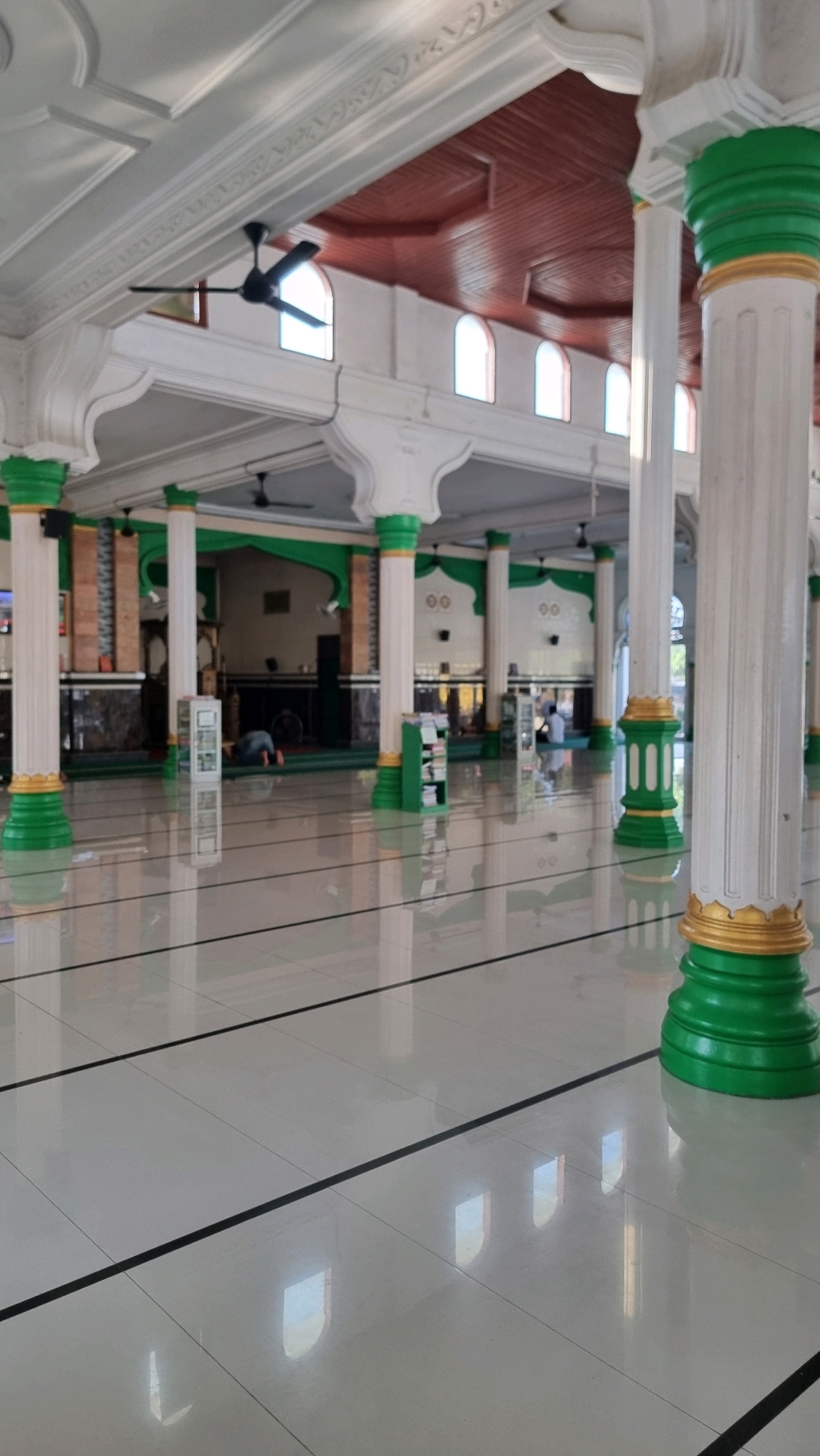 The image depicts the interior of a mosque in Blangrambong, Indonesia. The foreground is dominated by a highly polished, light-colored tiled floor with thin black lines marking sections. Several large, white, fluted columns, accented with green and gold bases, are strategically placed throughout the space. In the mid-ground, the mosque's prayer hall is visible, featuring green carpeted floors and more white columns with similar green and gold accents. Various objects are scattered in the prayer hall, including small bookshelves holding what appear to be books, and ceiling fans. Two individuals are present in the prayer hall; one is kneeling on the carpet in a prayer posture, while the other is seated. The ceiling is constructed of dark wood panels with a decorative pattern, and arched windows with translucent panes are visible along the upper walls, allowing natural light to filter in. The overall impression is one of cleanliness, spaciousness, and a serene environment dedicated to prayer.