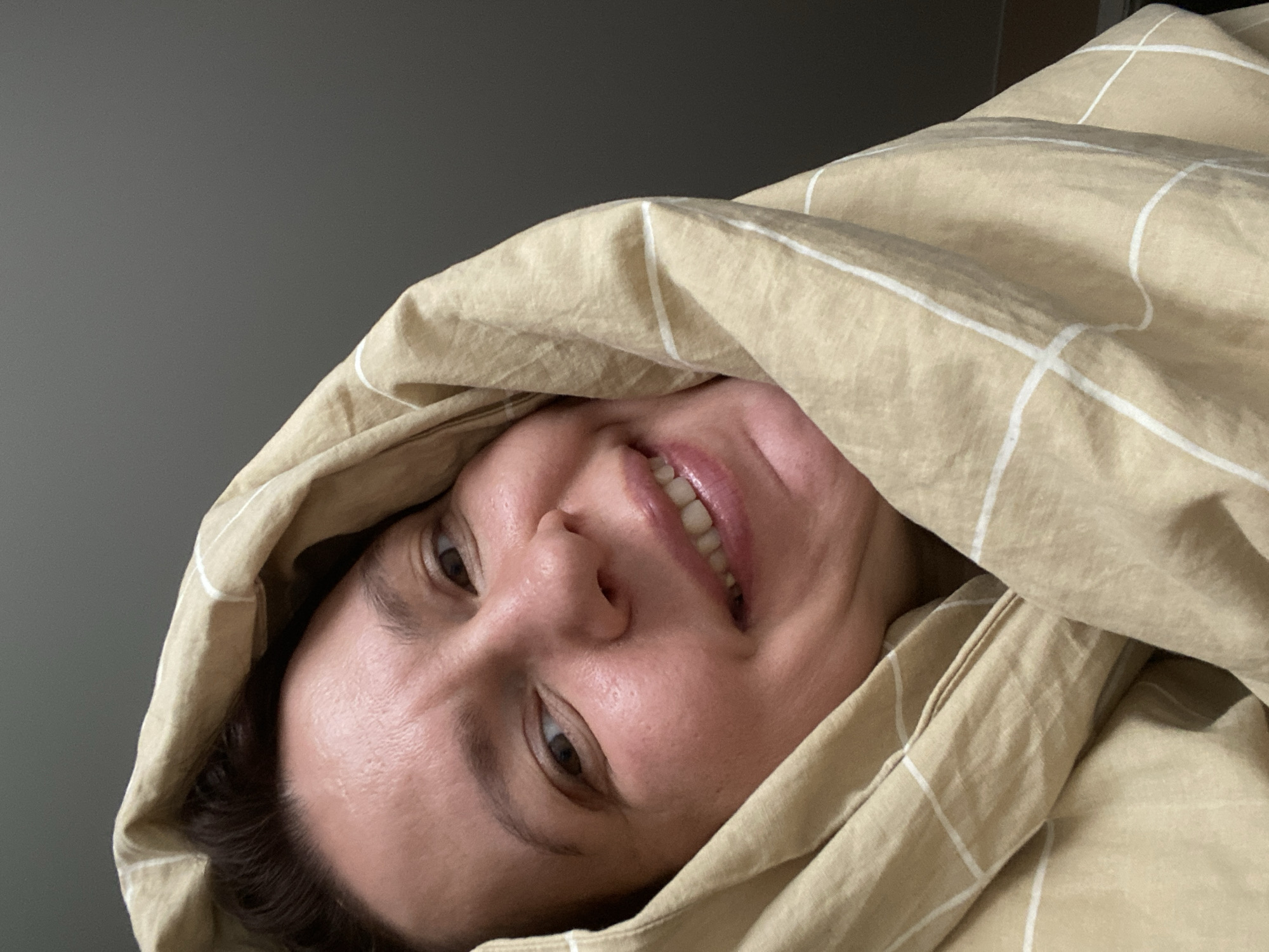 A woman is lying down, covered in a beige blanket with white stripes. She is smiling and looking at the camera. The background is a grey wall. There is no indication of the location being Chaikovsky, Russia.