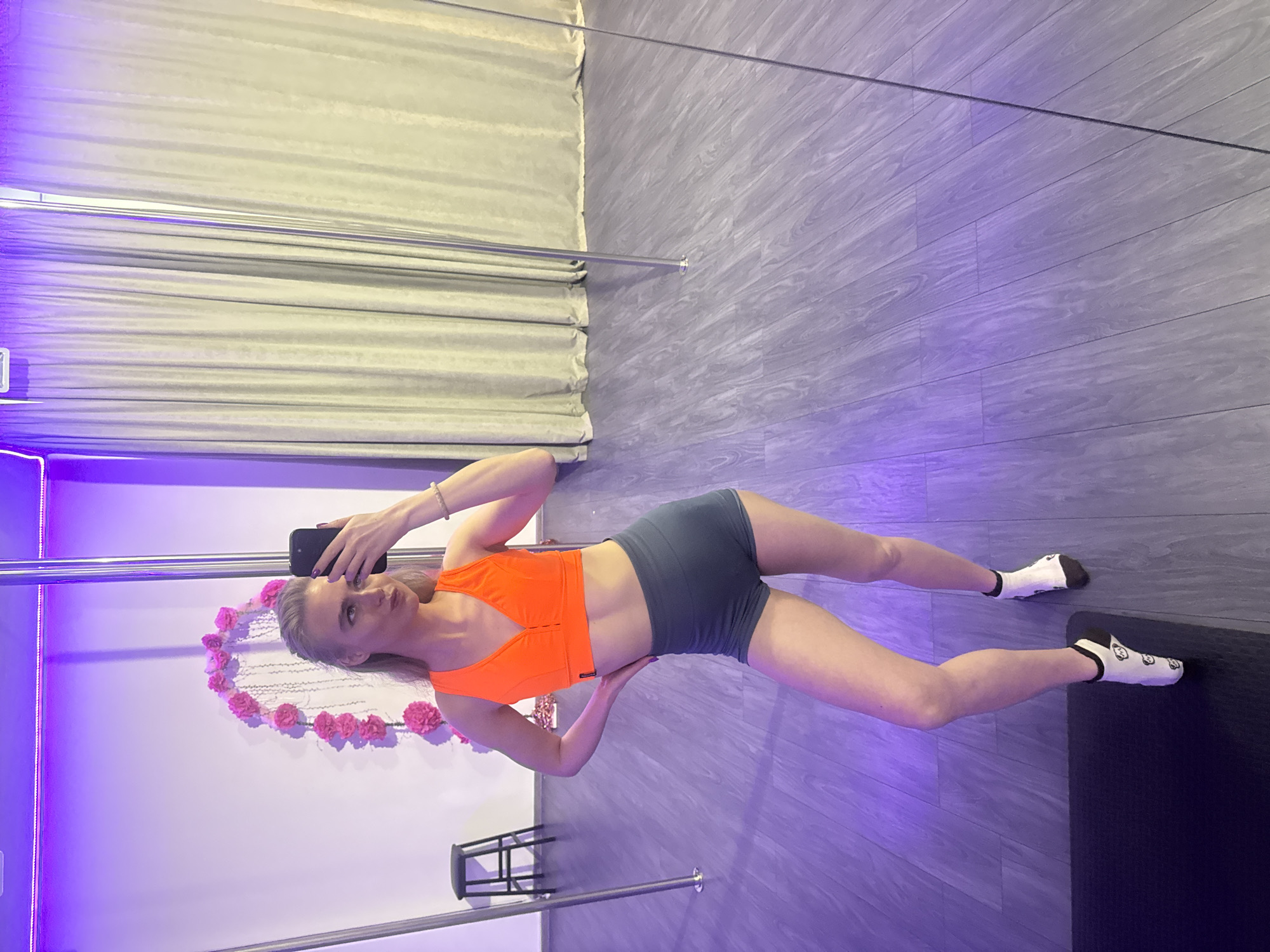 The image, rotated 90 degrees counter-clockwise, presents a full-body view of a woman posing in what appears to be a fitness or pole dance studio. She stands with her legs spread wide apart, holding a black smartphone in her right hand to take a mirror selfie, and looks directly at the camera with a neutral expression.
The woman has light-colored hair and is wearing a vibrant orange sports bra or crop top paired with dark grey shorts. She has white ankle socks with a dark heel and toe, and a subtle pattern. A metallic pole extends vertically on the left side of the frame, suggesting a pole dancing environment, and another pole base is visible towards the bottom.
The studio features a light grey floor with a wood-grain pattern, and the walls are predominantly white, illuminated by a distinct purple ambient light. A heart-shaped decoration made of pink flowers or roses adorns the white wall behind the woman. Light-colored curtains or blinds are visible in the background on the far left. A small, dark stool or chair is positioned behind her left leg, and a dark exercise mat lies on the floor to the right. No visible text is clearly discernible in the image.