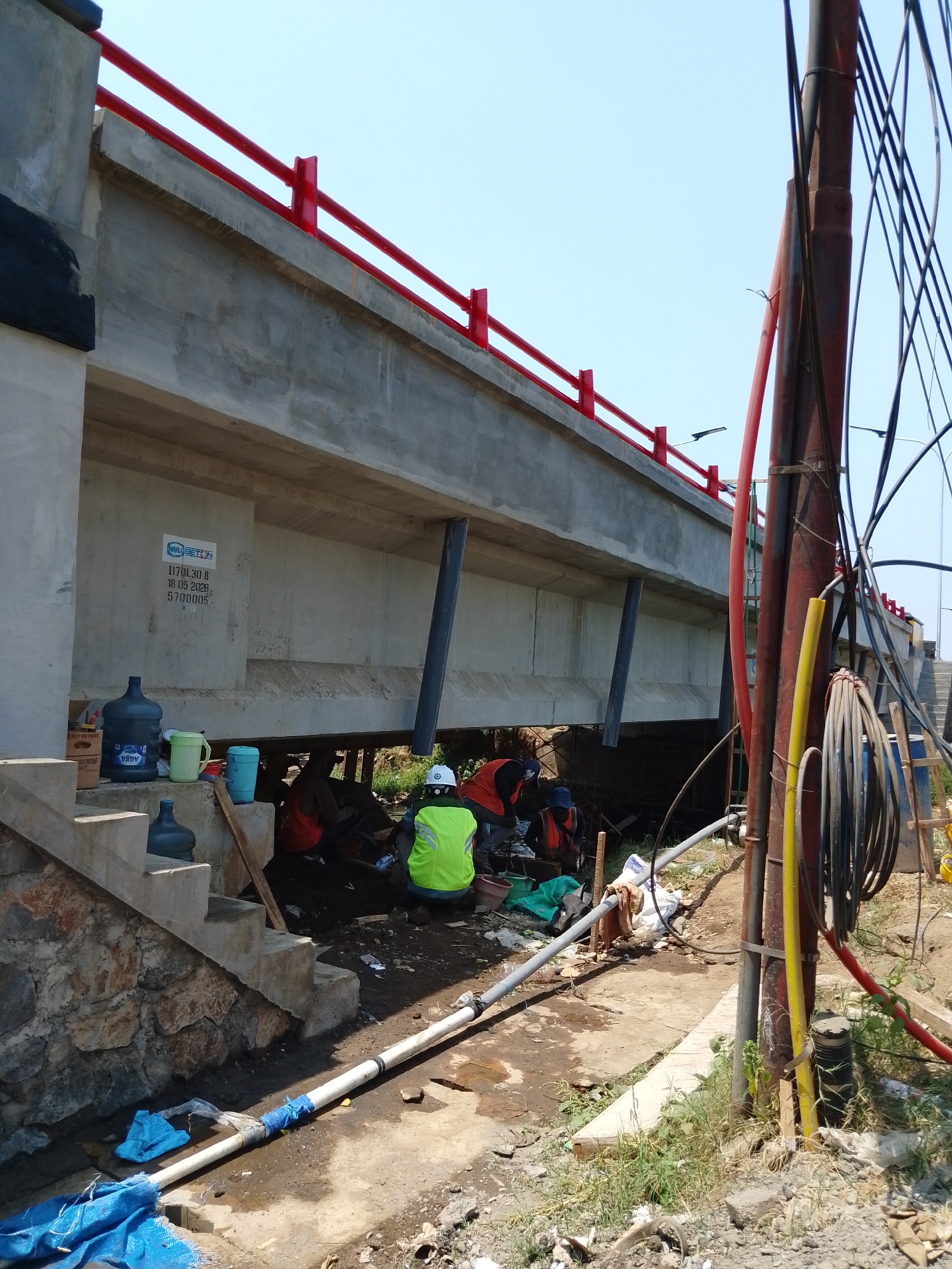 In Pasuruan, Indonesia, under a bridge under construction, a group of men are engaged in work. One man, wearing a bright green vest and a white hard hat, is seated. Another man, wearing a blue cap and an orange vest, is kneeling, possibly working on a pipe that runs along the ground. A third man, also in an orange vest, is partially obscured. The environment is rough, with dirt, debris, and construction materials scattered around. A large concrete beam forms the underside of the bridge, supported by angled pillars. Red railings line the top of the bridge, and electrical wires and conduits are visible on a pole to the right. Several large water jugs and cups are placed on concrete steps to the left of the scene.