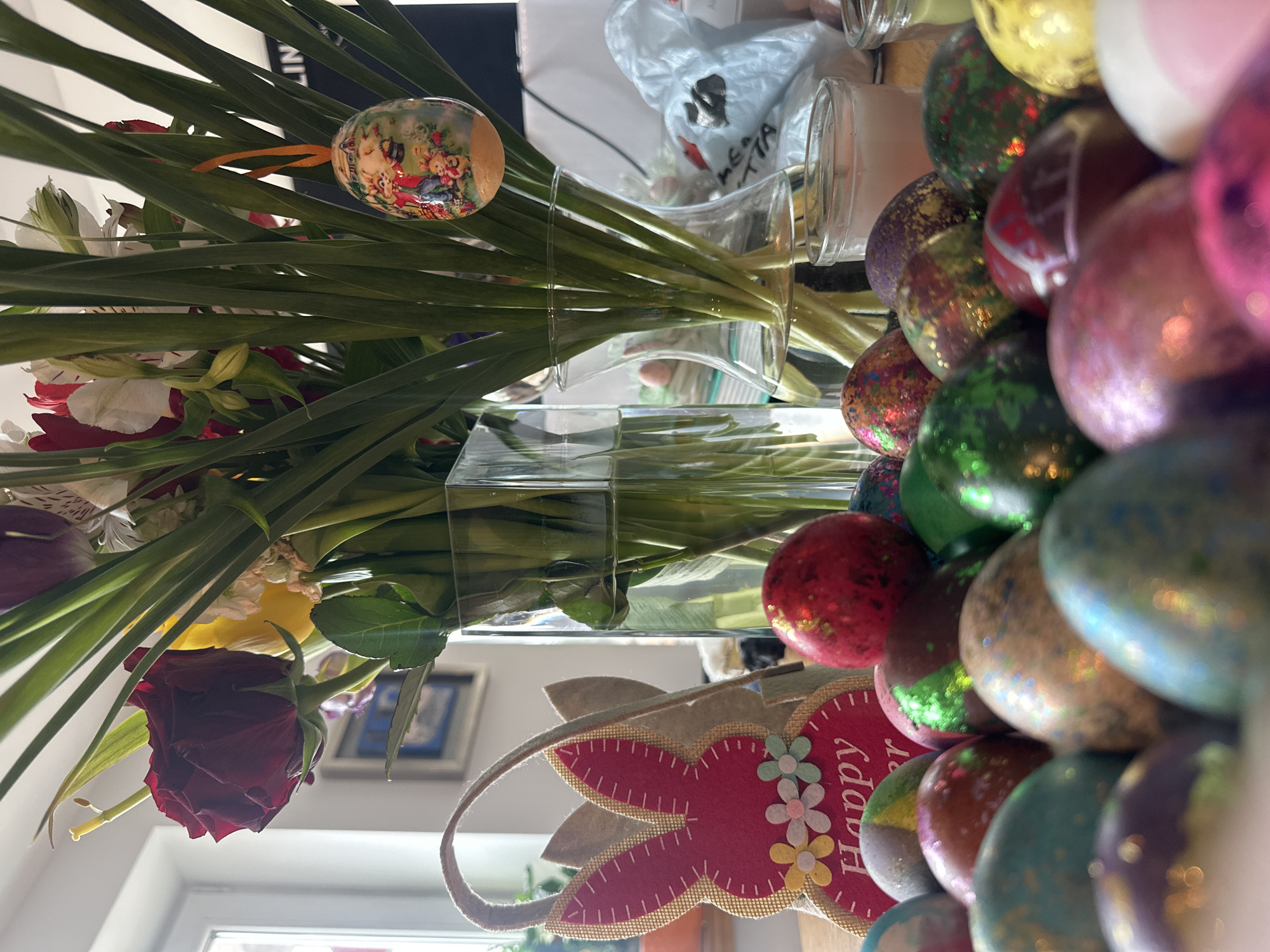 The image displays a still life arrangement with various objects, primarily focused on Easter and spring themes. A bouquet of flowers, including roses, lilies, and other assorted blooms, is arranged in a clear glass vase. An Easter egg, decorated with a pastoral scene, hangs among the flower stems. To the right of the flowers, a collection of Easter eggs, covered in glitter and various colors, are stacked. A felt Easter bunny decoration with the word "Happy" on it is visible near the base of the stack of eggs. The items are arranged on a reflective surface, likely a tabletop.