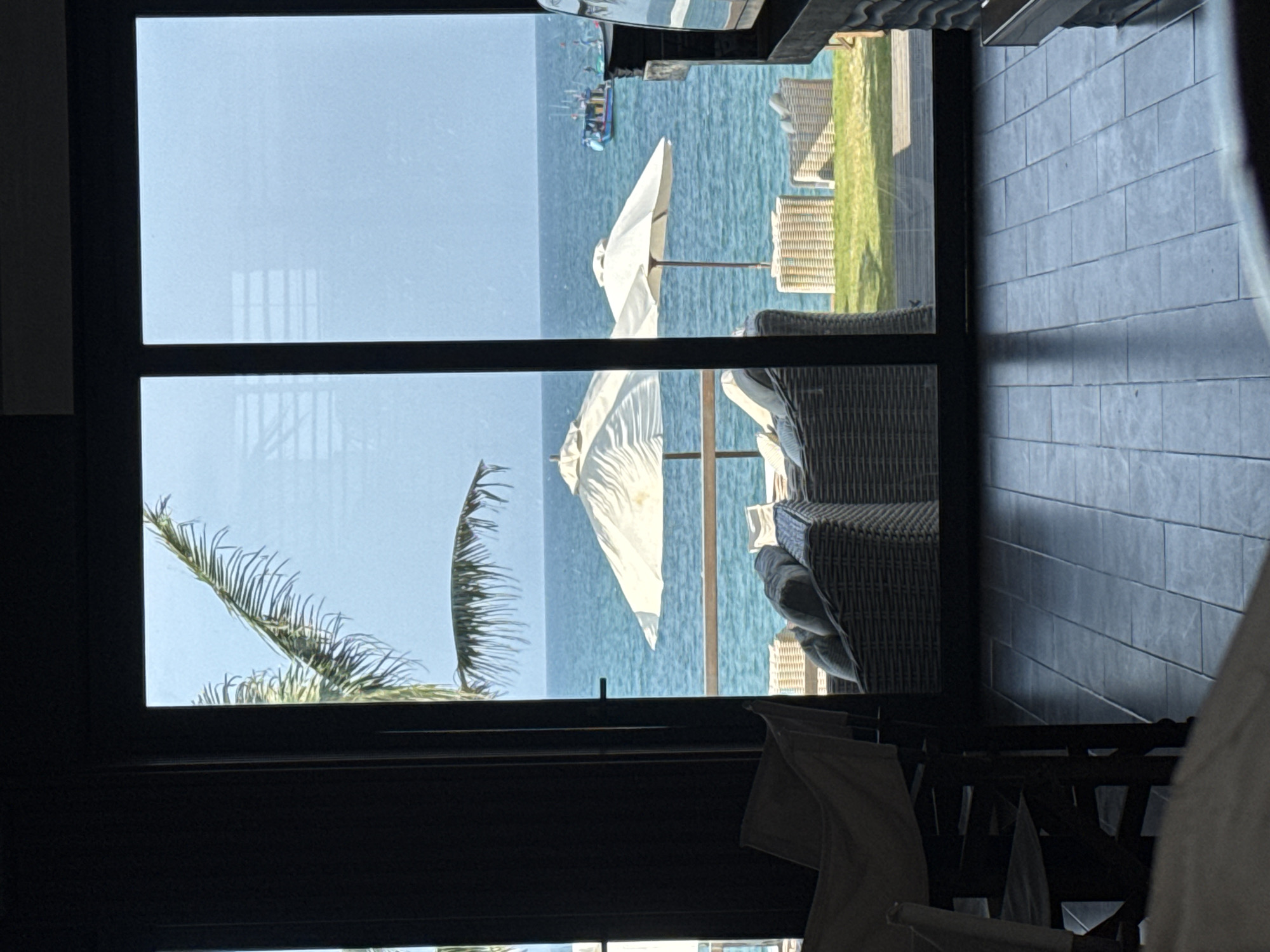 View from inside a building looking out to the beach. Two white umbrellas are visible on the beach along with several chairs. There is a group of people on a boat in the water. A palm tree is in the lower left of the frame. The beach is located near Đà Nẵng, Vietnam.