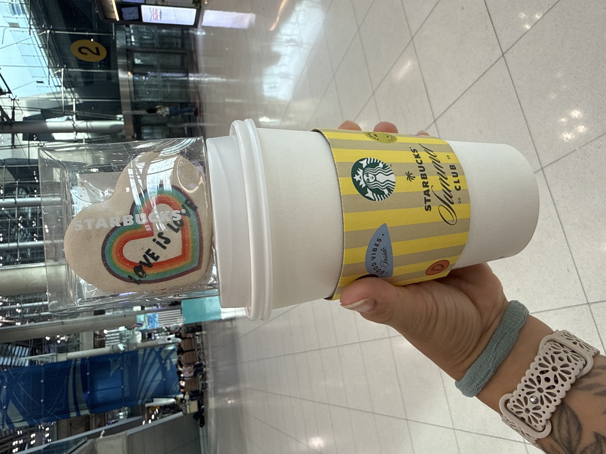 A person's hand is holding a Starbucks paper cup with a yellow and white striped sleeve. On the sleeve, the Starbucks logo and the words "Summer CLUB 2020" are visible. Above the cup, a heart-shaped cookie decorated with a rainbow and the words "LOVE IS LOVE" in black lettering is encased in clear plastic packaging. The background suggests an airport terminal in the city of Nong Prue Subdistrict, Thailand. Large pillars, a blue banner with Thai text, glass doors, and seating areas with people are visible. The floor is tiled with a grid pattern.