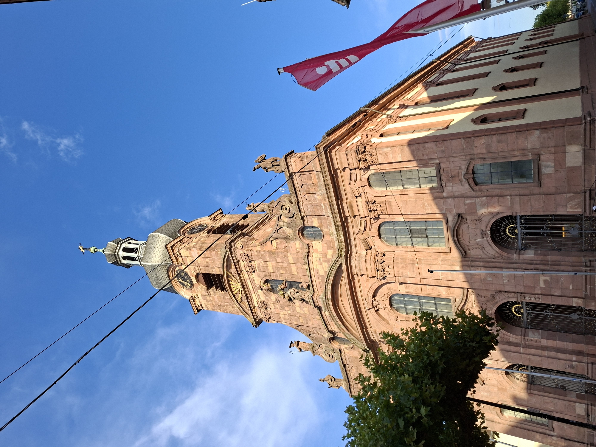 This is a low-angle, eye-level, medium shot of the exterior of the Church of the Ascension (Aufkirche) in Rastatt, Germany, on a clear, sunny day. The church is constructed primarily of reddish-brown sandstone and features a prominent bell tower with a dark, multi-tiered spire topped with a cross. The tower has clock faces and arched openings. The facade of the church is ornate, with statues of figures in niches, decorative moldings, and arched windows with intricate metalwork on the ground level.

To the left of the church, a large, leafy green tree casts a shadow onto the building. To the right, two bright red flags with a white logo are prominently displayed, fluttering in the breeze. The flags appear to be advertising something. Overhead, black utility lines stretch across the bright blue sky, which has a few wispy white clouds. The side of the church visible in the background is painted a light color with regularly spaced, smaller windows. The overall scene suggests a peaceful urban environment during daylight hours.