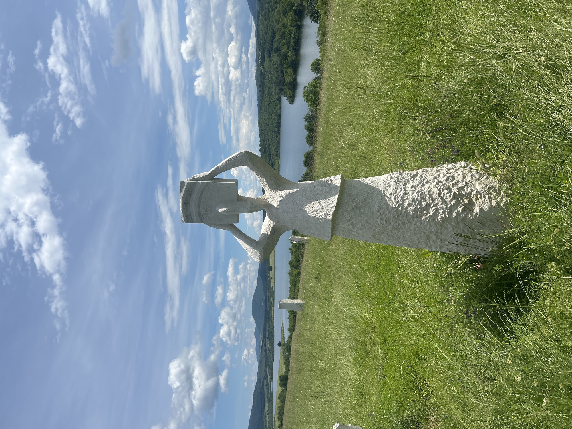The image displays a large, light-colored stone sculpture set against a scenic natural backdrop. The sculpture, crafted from a textured, chiseled material, depicts a highly stylized human figure, possibly female, holding a rectangular object or book above its head. The figure's form is abstract and geometric.
The sculpture is positioned on a vibrant green hillside covered in tall grass and subtle wildflowers. Beyond the immediate foreground, a calm body of water, likely a lake or reservoir, spans the midground, reflecting the clear sky. On the far side of the water, a series of forested hills and mountains rises, their slopes densely covered in trees. The sky overhead is bright blue with scattered white clouds, including both wispy cirrus and fluffy cumulus formations.
The scene is captured on a clear, sunny day, indicated by the strong light and distinct shadows cast by the sculpture. The setting evokes a sense of tranquility and natural beauty, suggesting an outdoor art installation or sculpture park integrated with the landscape. There is no discernible activity taking place other than the static presence of the sculpture within its environment. No text is visible in the image. The location is Gardevtsi, Bulgaria.