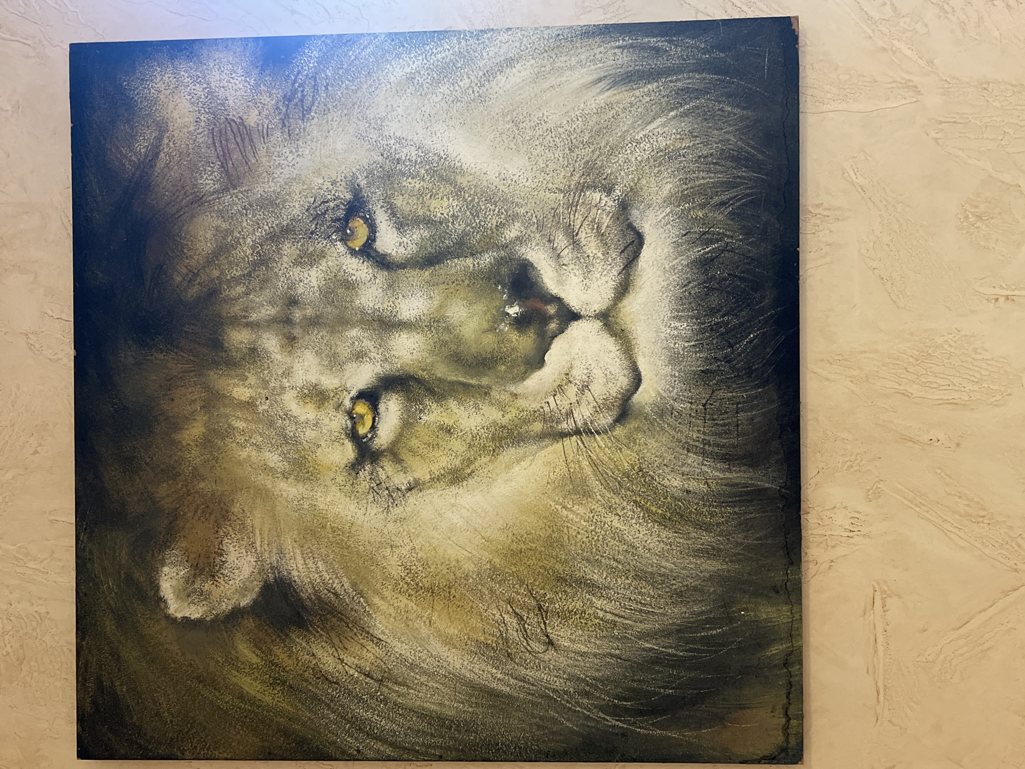 A close-up, slightly angled photograph captures a painting of a lion's face on a textured, light beige wall. The painting itself is a detailed depiction of a lion, rendered in muted tones of gold, brown, and grey. The lion's piercing yellow eyes are a focal point, conveying a sense of intensity. Its mane is depicted with individual strands, creating a textured effect that blends into the dark background. The painting appears to be framed with dark wood, and the bottom edge shows some wear and tear, revealing a lighter material underneath. The wall behind the painting has a rough, stucco-like texture with visible vertical and horizontal indentations. The lighting in the scene appears to be coming from the right side, casting a slight sheen on the painting and creating subtle shadows on the wall. There are no people or animals present in the photograph, only the painting and the wall it hangs on. The image does not contain any information about its location in the city of Bender, Moldova.